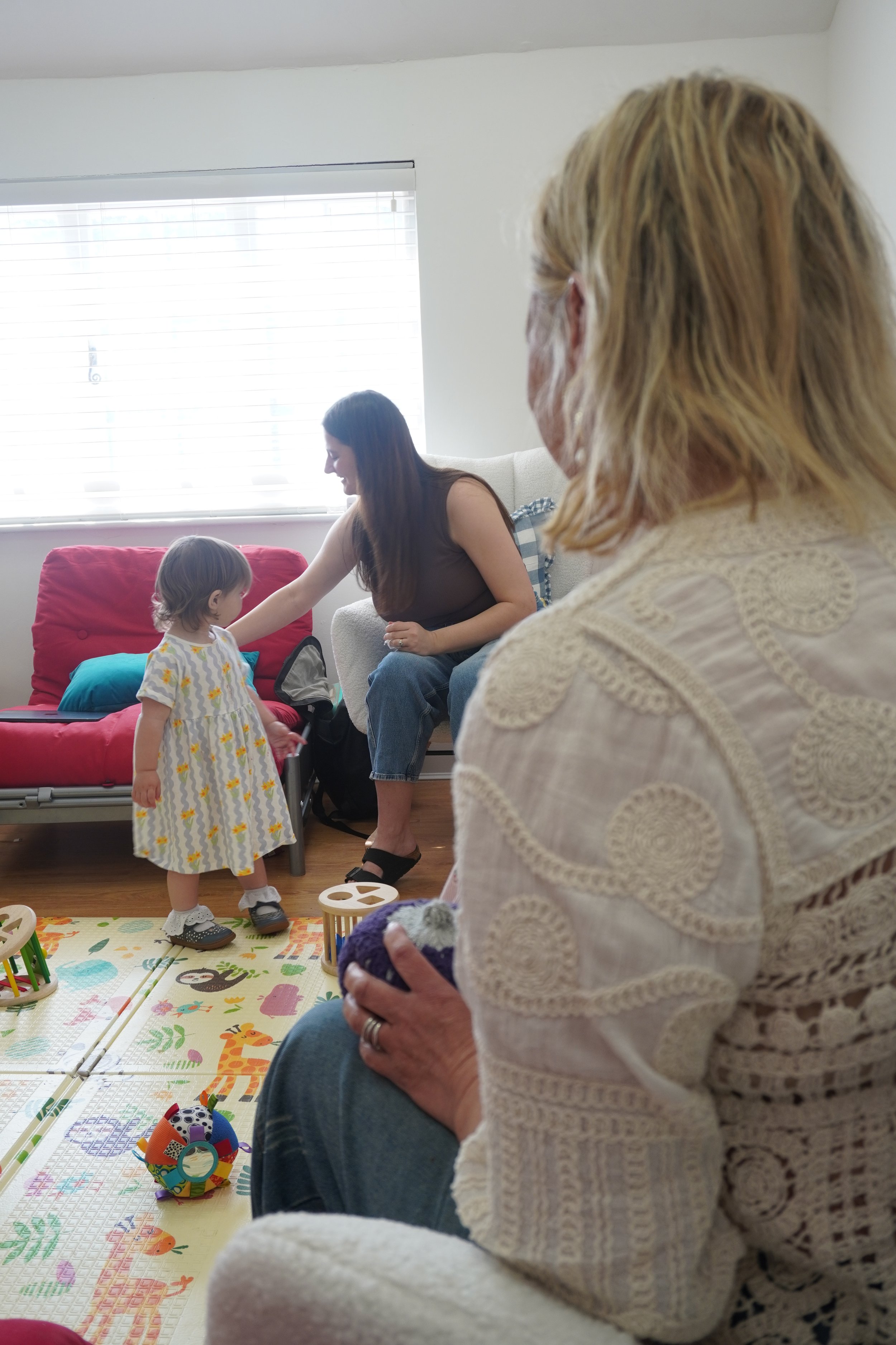 A family gathering indoors, with a young girl in a patterned dress standing next to family members sitting on a colorful mat with toys, a woman interacting with the girl, and an older woman with blonde hair holding a colorful ball.
