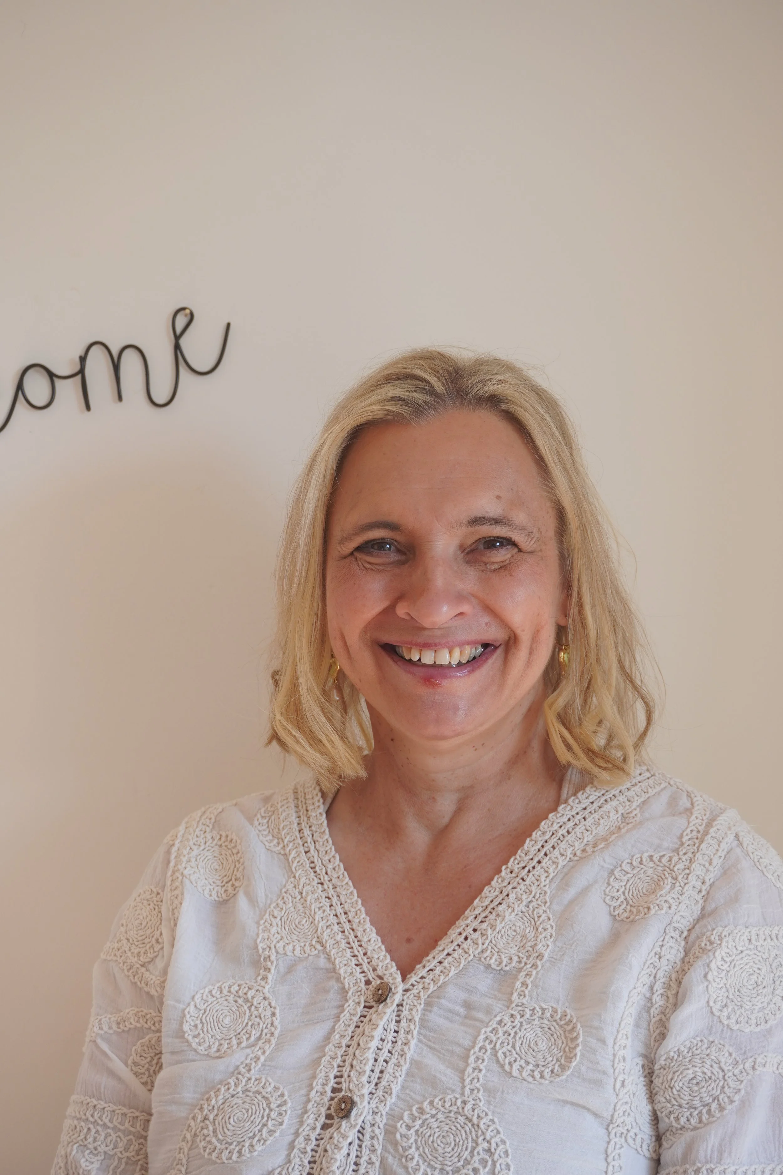 A woman with shoulder-length blonde hair, smiling, wearing a cream-colored embroidered blouse, standing in front of a beige wall with a decorative metal sign partially visible in the background.
