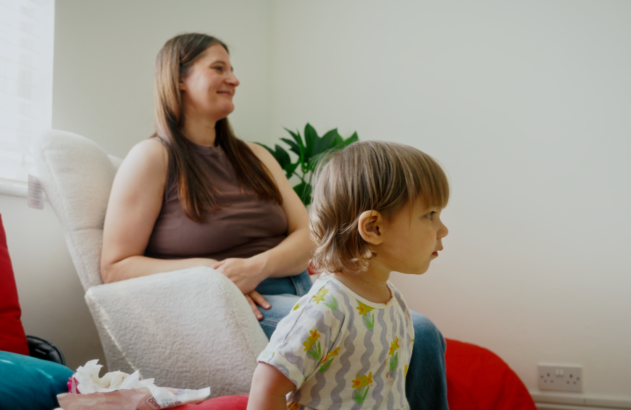 A woman and a young girl sitting and smiling in a bright room, with a plant in the background.
