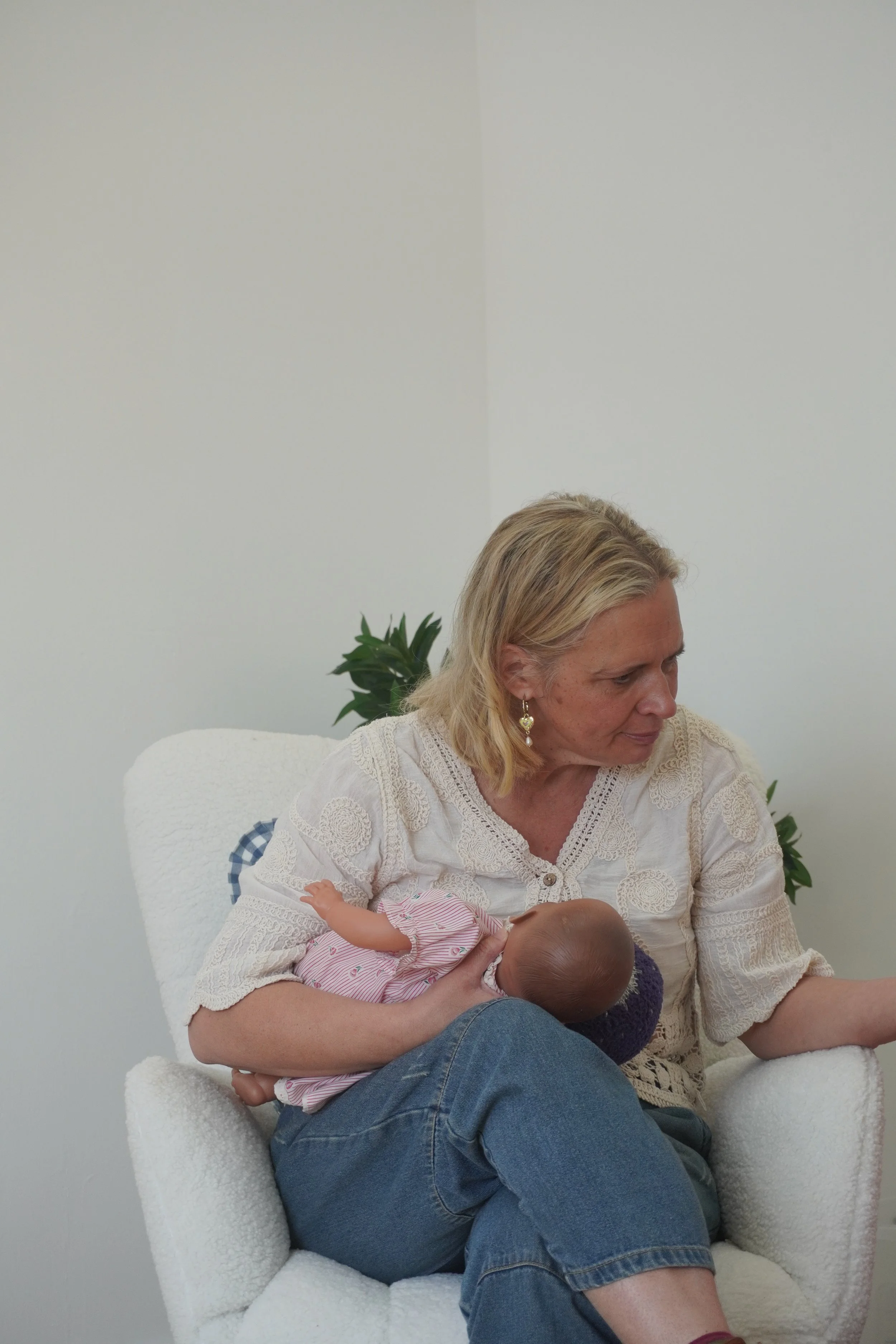 A woman sitting on a white chair, holding a sleeping baby in her arms, with a potted plant behind her and a white wall in the background.
