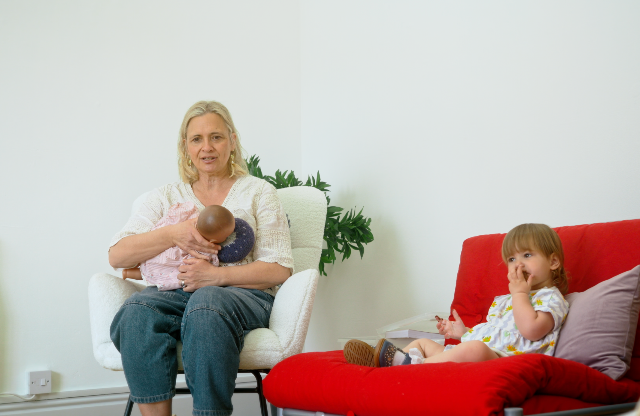 An older woman with blonde hair holding a baby on her lap, sitting on a white armchair in a living room. A young girl with curly red hair, sitting on a red couch, is listening and looking thoughtfully. There are plants in the background and a plain white wall.