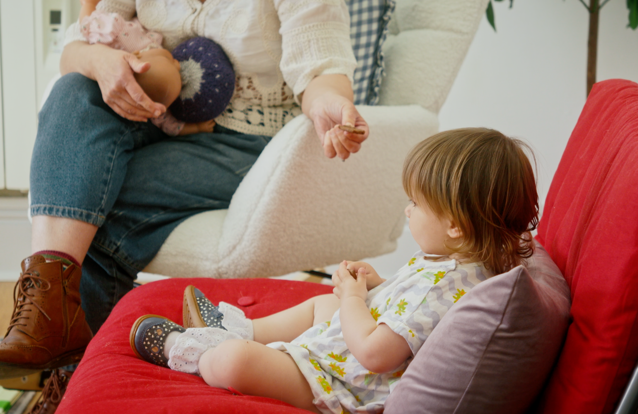 A young girl with brown hair sitting on a red couch, wearing a white dress with a colorful floral pattern and black shoes with white polka dots, looking at a woman holding a baby. The woman, sitting on the couch, wears jeans, a cream sweater, and brown boots, holding a baby wearing pink and white striped pajamas and a purple knit hat with white stars. Part of a white armchair and a green plant are visible in the background.