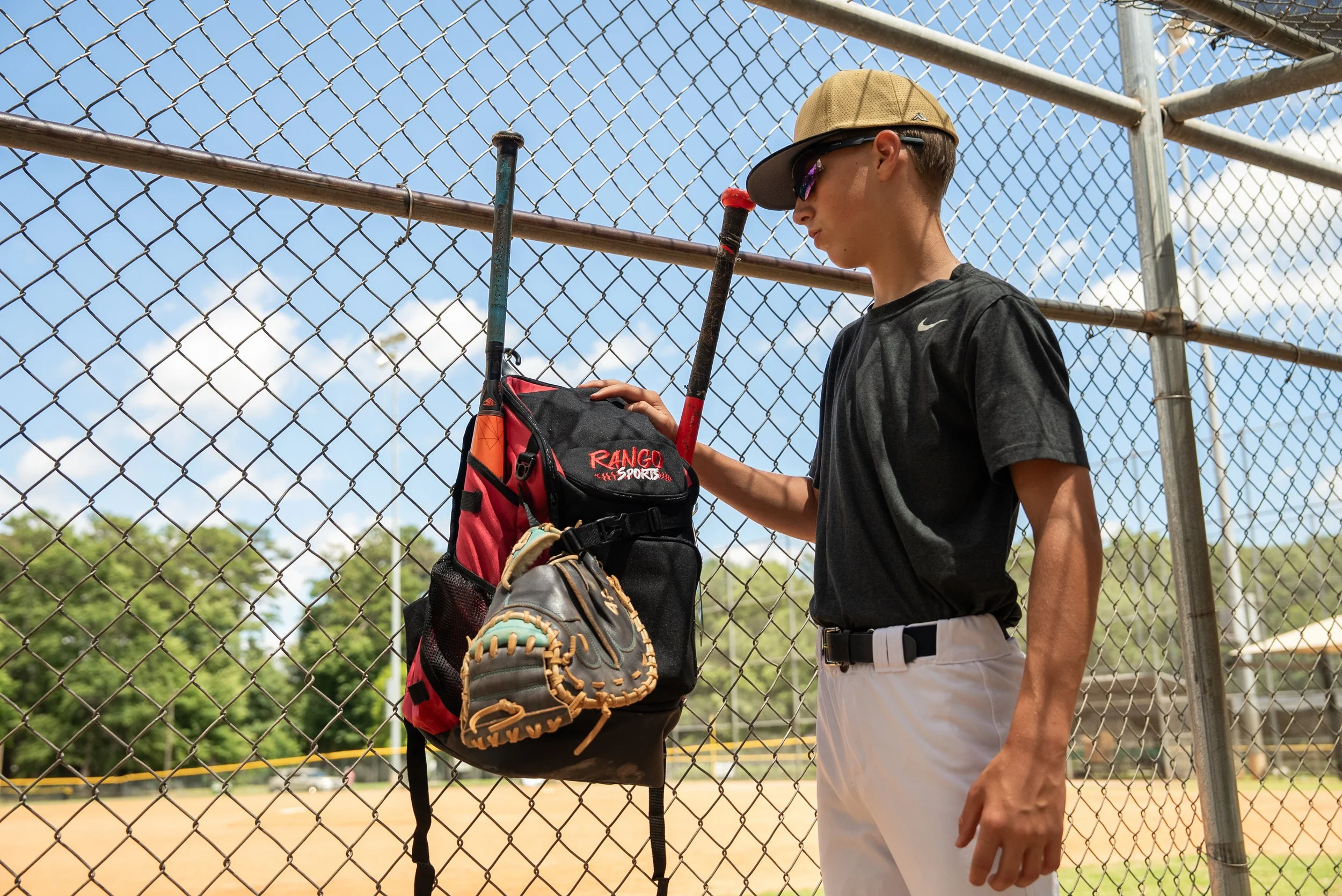 A young baseball player stands behind a chain-link fence with his baseball glove, backpack, and bat, wearing sunglasses and a tan cap, during a sunny day at the baseball field.
