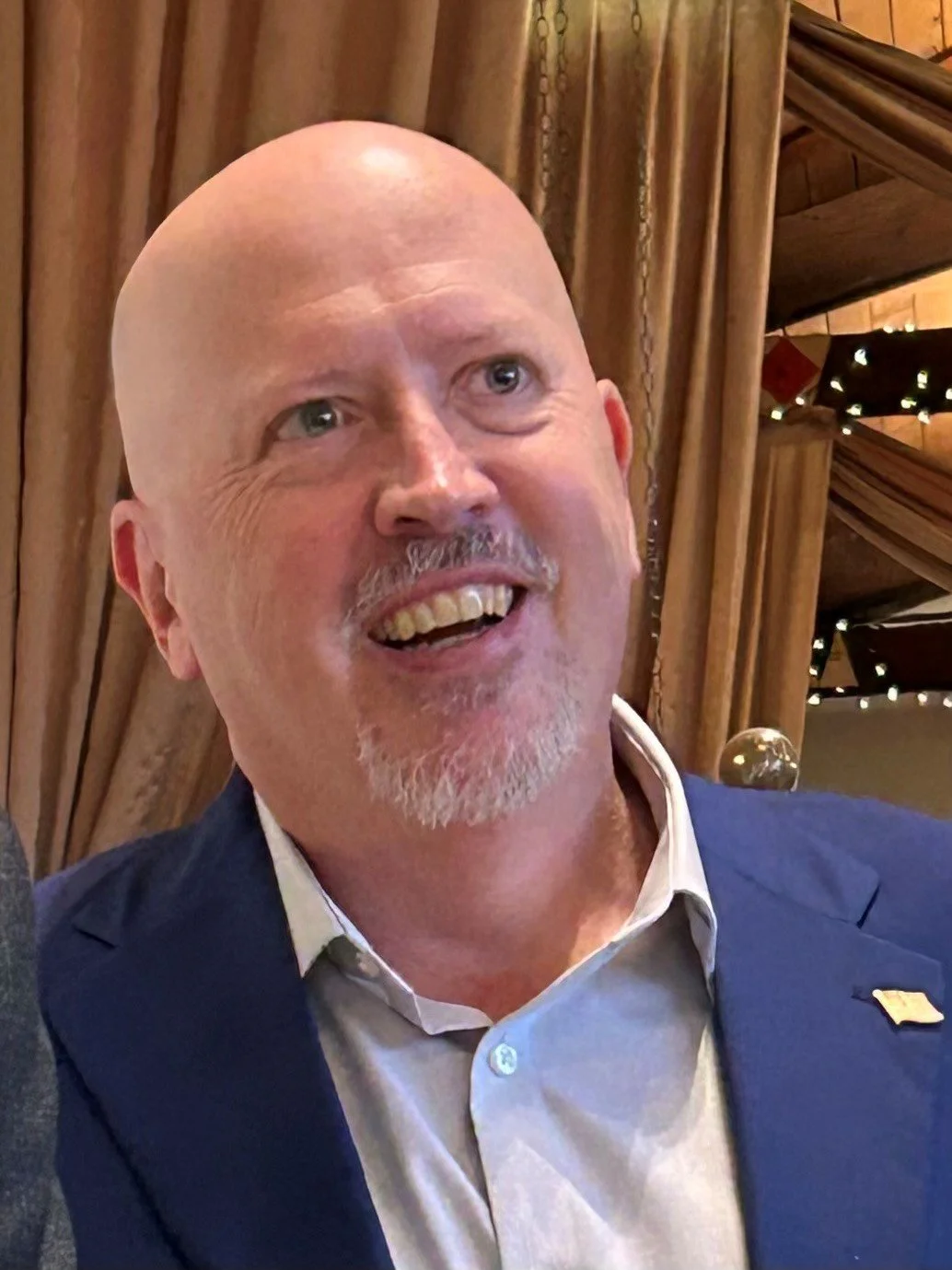 A smiling middle-aged man with a bald head, gray goatee, light-colored eyes, wearing a navy blazer and white dress shirt, sits in front of wooden wall panels and curtain with string lights in the background.