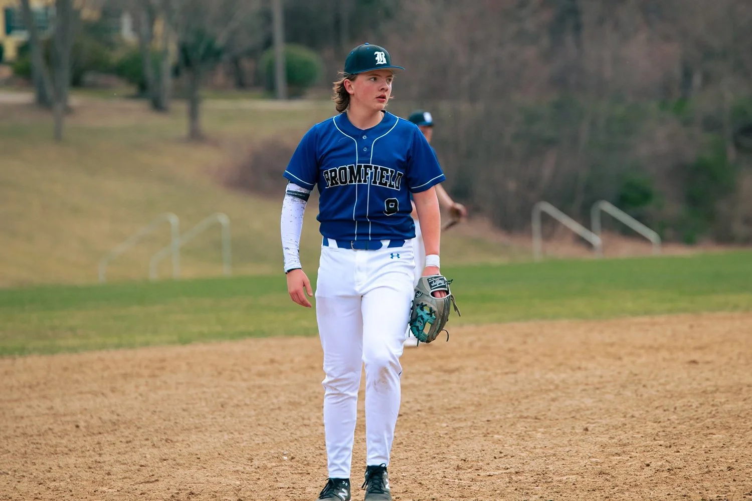 A young baseball player standing on the field, wearing a blue jersey with 'PROMFIED' and the number 9, white pants, a baseball glove, and a black helmet.
