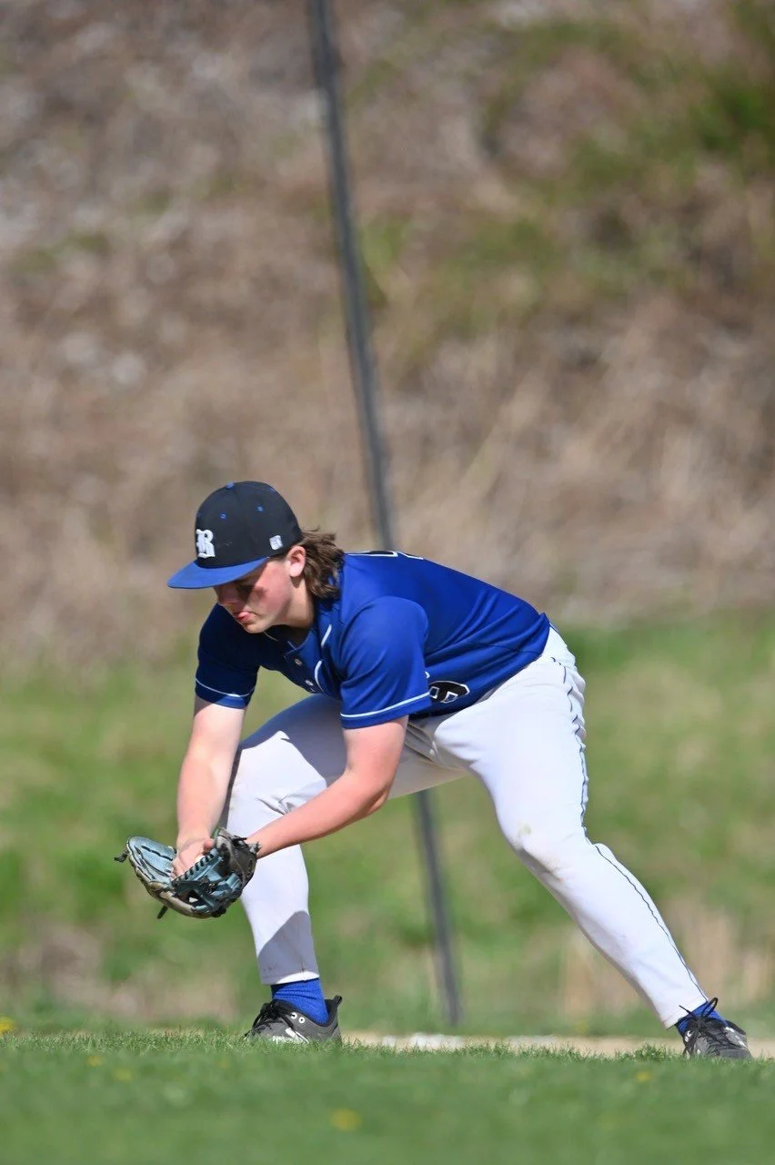 A baseball player in a blue jersey and white pants, wearing a black and blue cap, in a field, catching a ground ball with his glove.