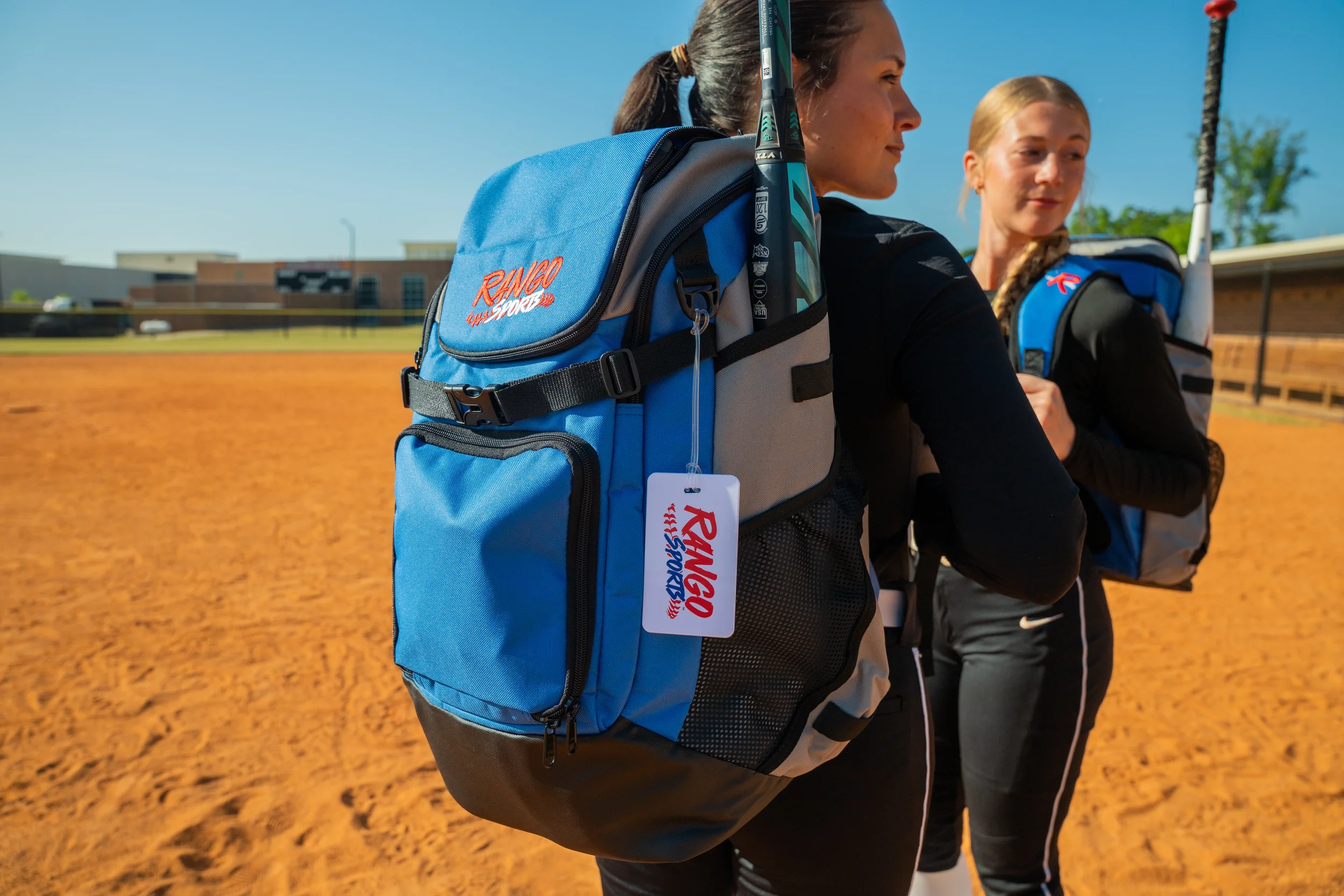 Two women are standing on a softball field, each carrying blue sports backpacks with the logo 'Ringo Sports' and walking side by side. The scene is outdoors with a clear blue sky and a school building in the background.