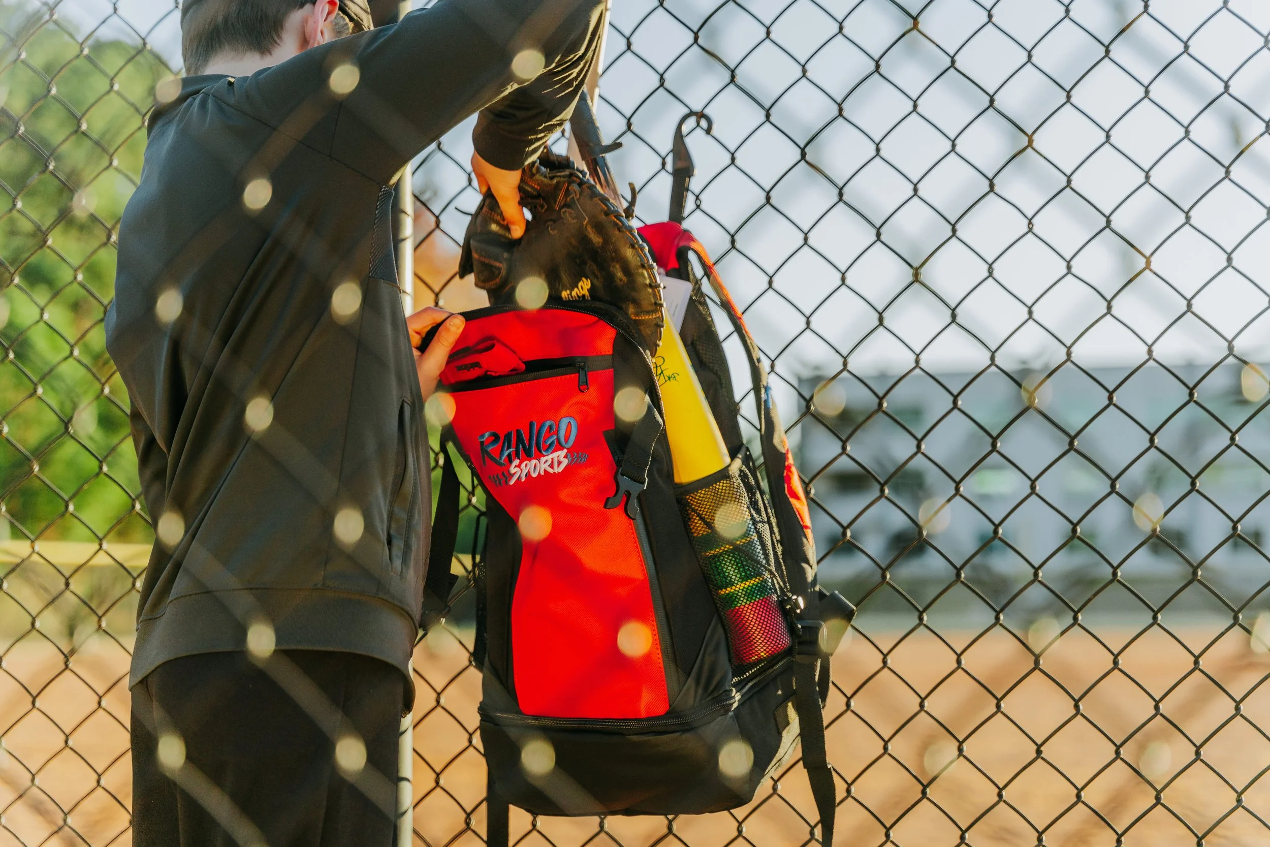 A person putting on a glove outside near a chain-link fence, with a sports bag hanging on the fence.