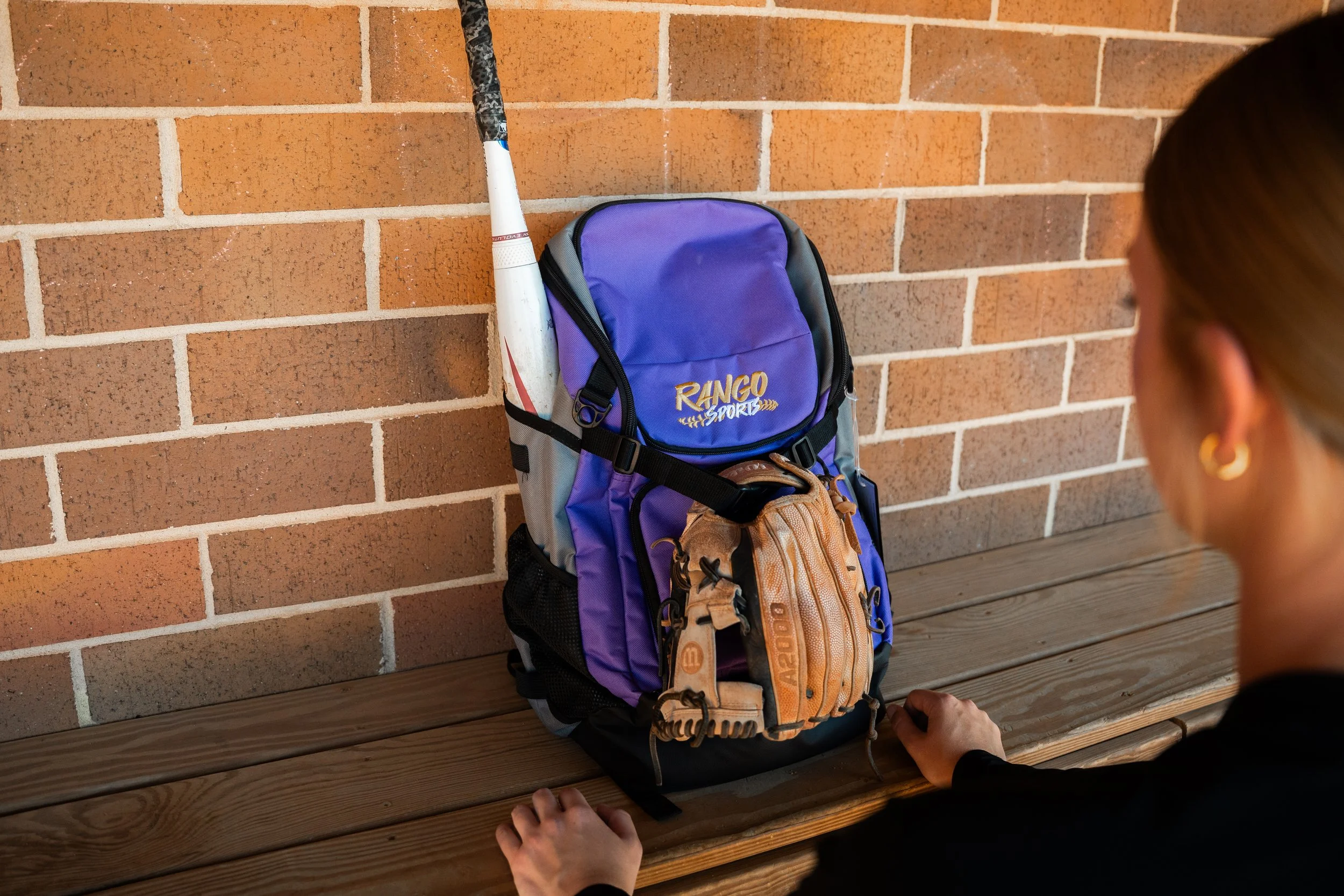 A purple RANGO sports backpack with a white baseball bat and a tan baseball glove hanging on the outside, placed on a wooden bench against a brick wall. A person with earrings is sitting nearby.