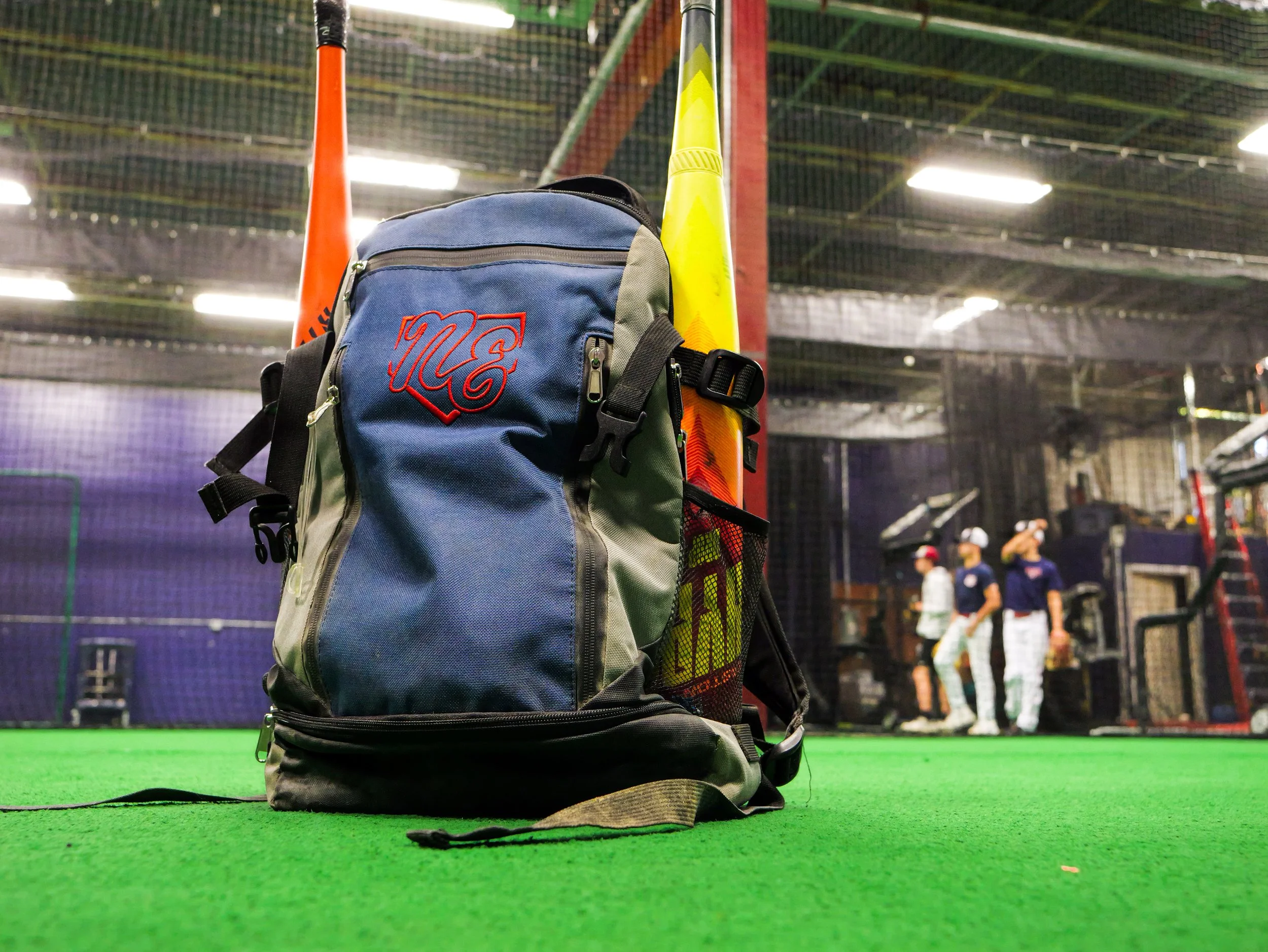 A backpack with a red logo on it, placed on the floor of an indoor sports facility with an artificial green turf. Behind the backpack, there are two traffic cones, one orange and one yellow, and a netted area where people can be seen practicing baseball or softball.