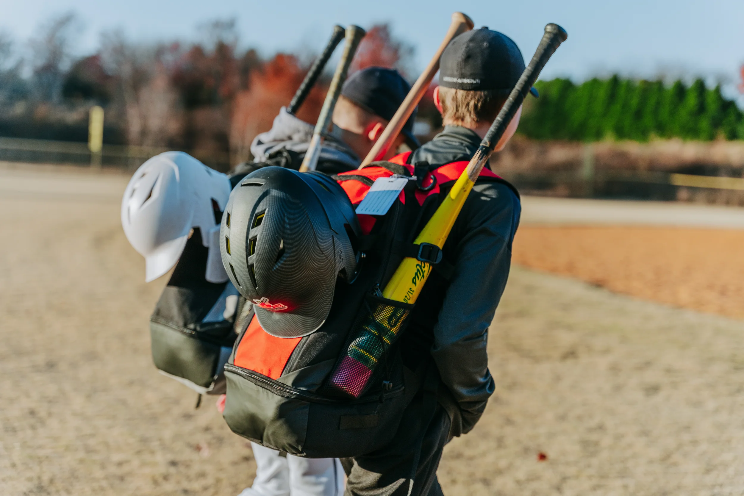 Close-up of two baseball players walking away, carrying bats, helmets, and gear on their backs, in a field with trees in the background.