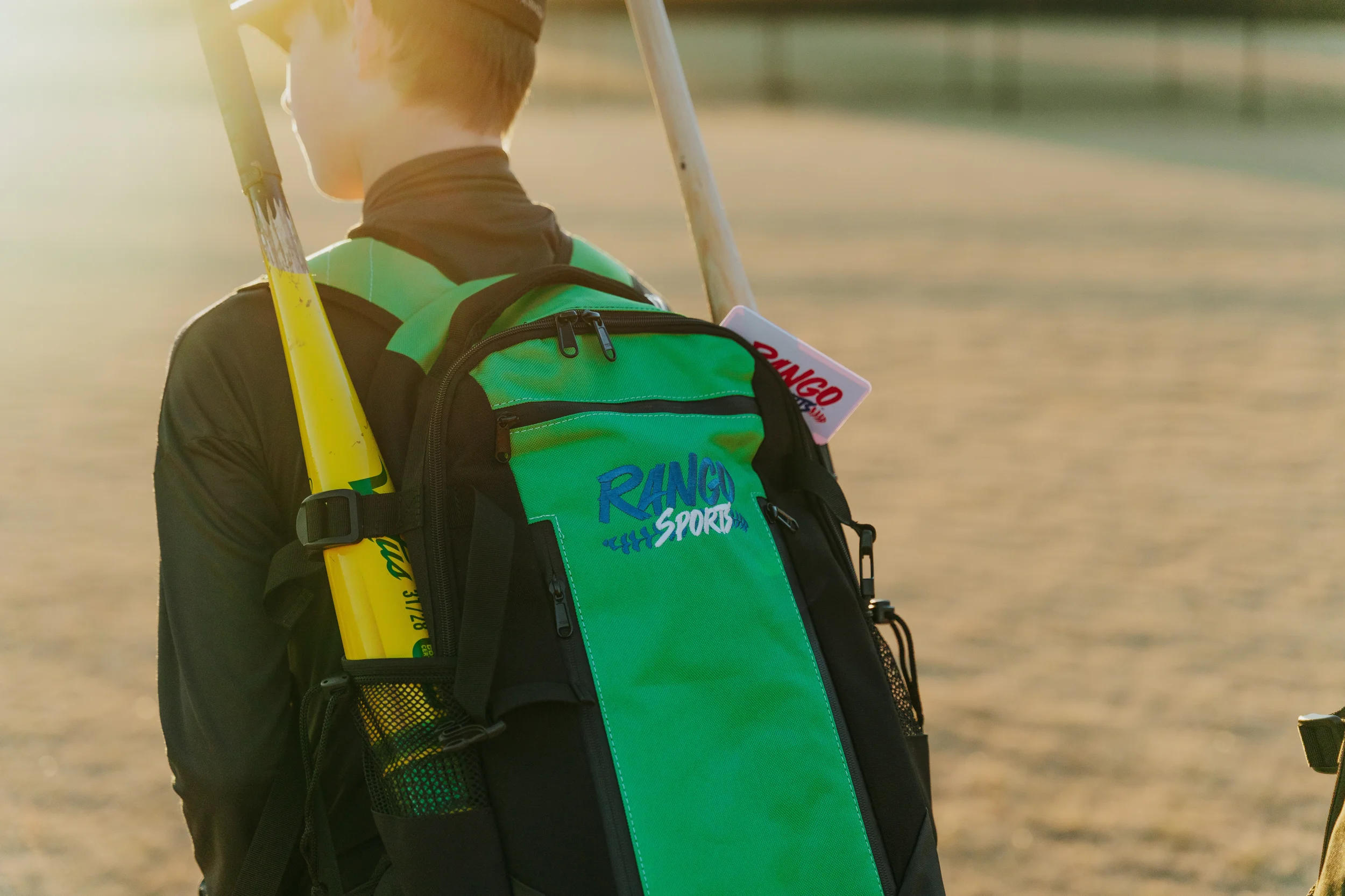 A person wearing a green backpack with the words "RANGO SPORTS" on it, standing outdoors in sunlight, holding a yellow bat, with a tag attached to the backpack.
