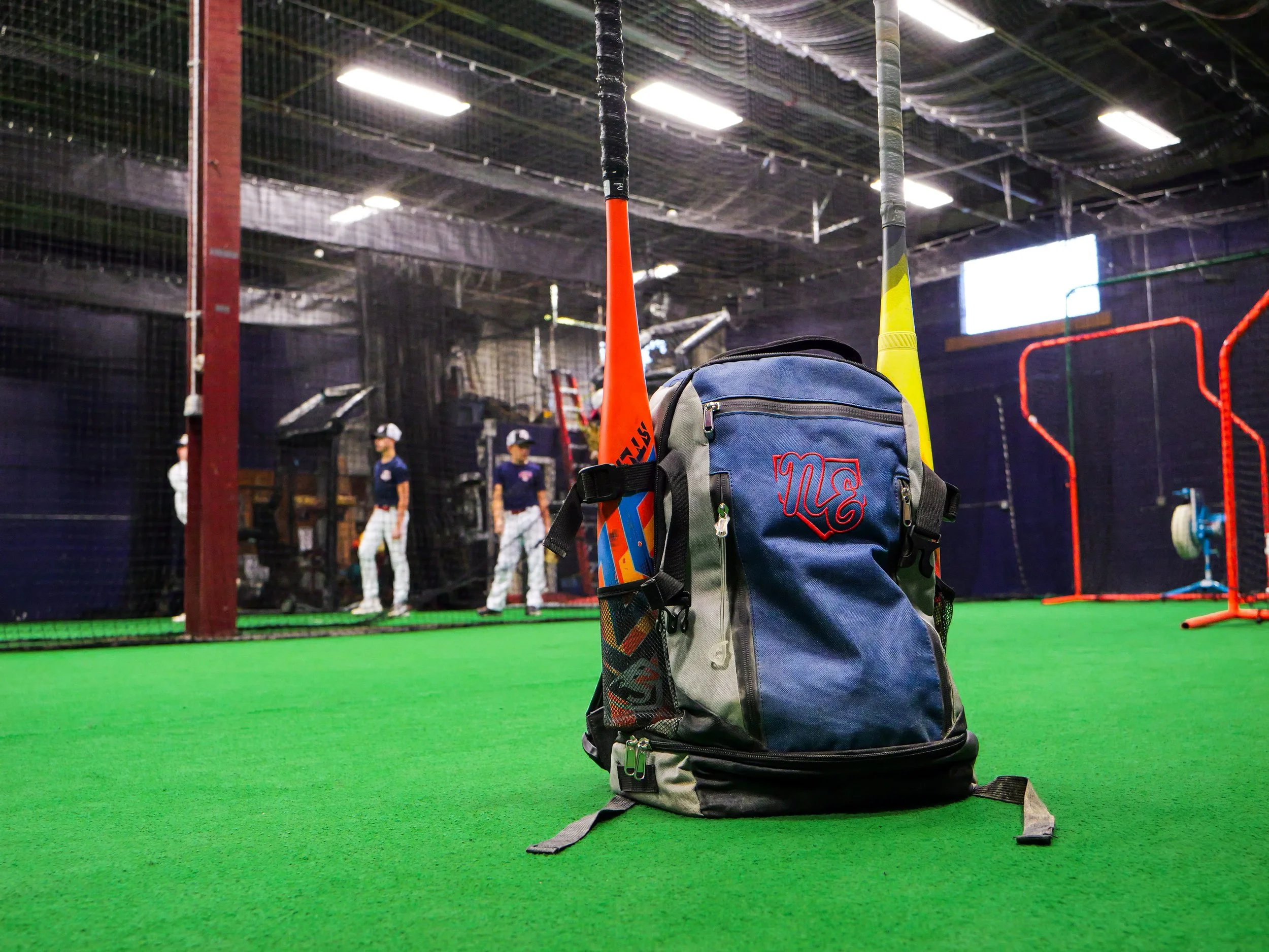 Backpack with a baseball bat and a water bottle placed on an artificial turf field, with people and sports equipment visible in the background of an indoor sports facility.