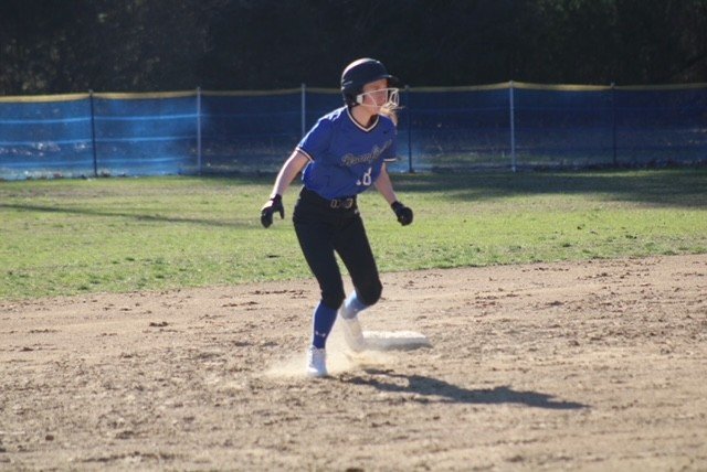 A female softball player in a blue uniform, helmet, and gloves running on a dirt field near first base during a game.