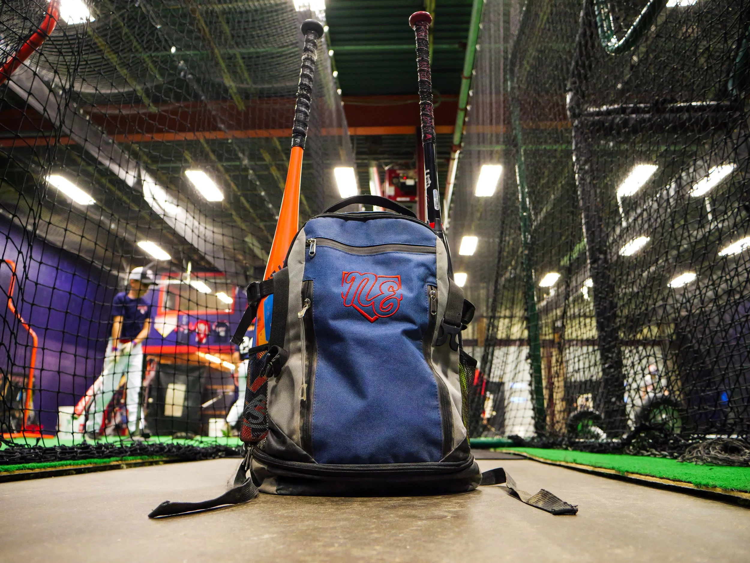 A backpack on the floor in front of a tennis practice cage with a person practicing tennis in the background.