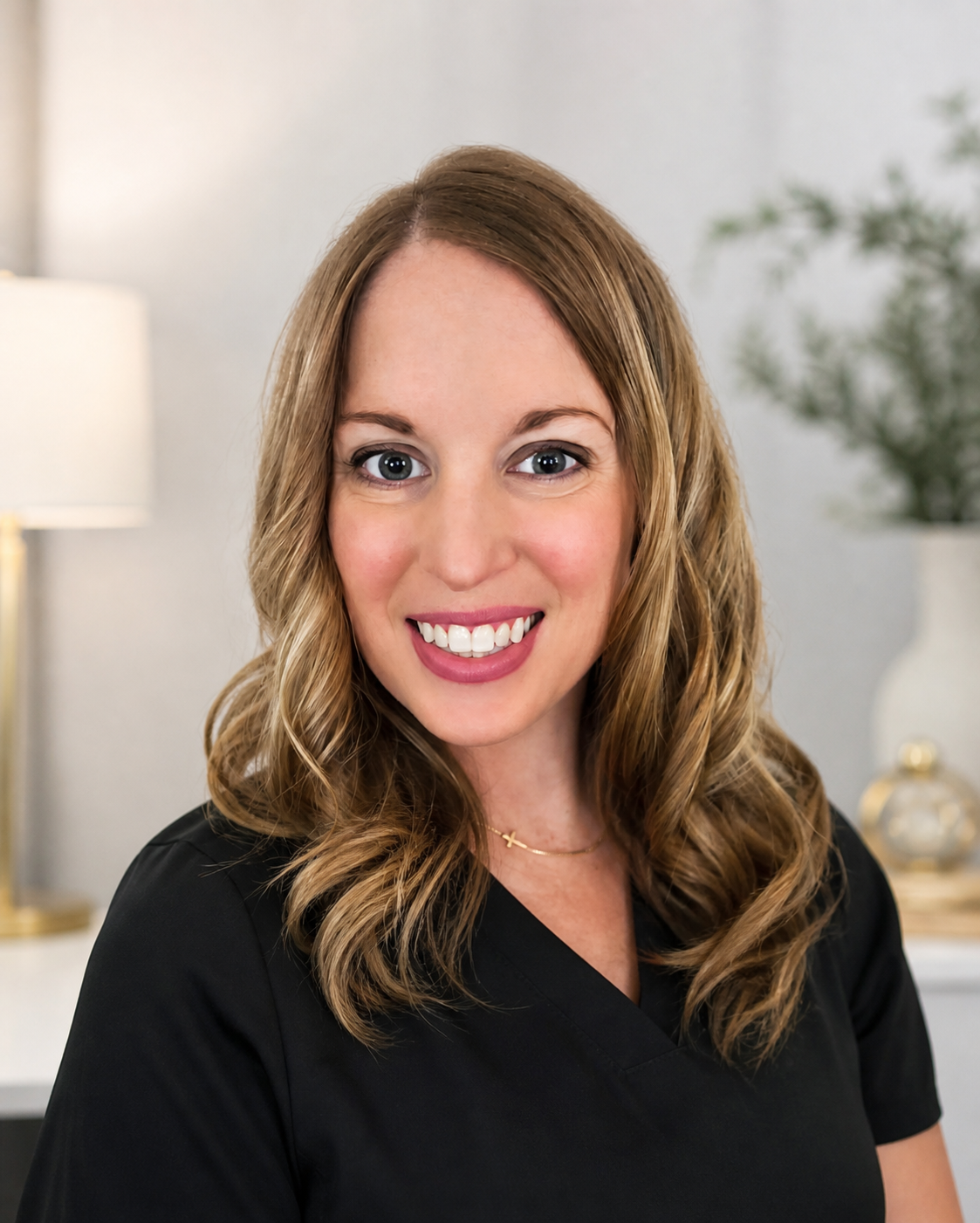 A woman with wavy light brown hair smiling in a room with a lamp, a plant, and decor in the background. Med Spa in Fishers, IN | B Beauty Aesthetics