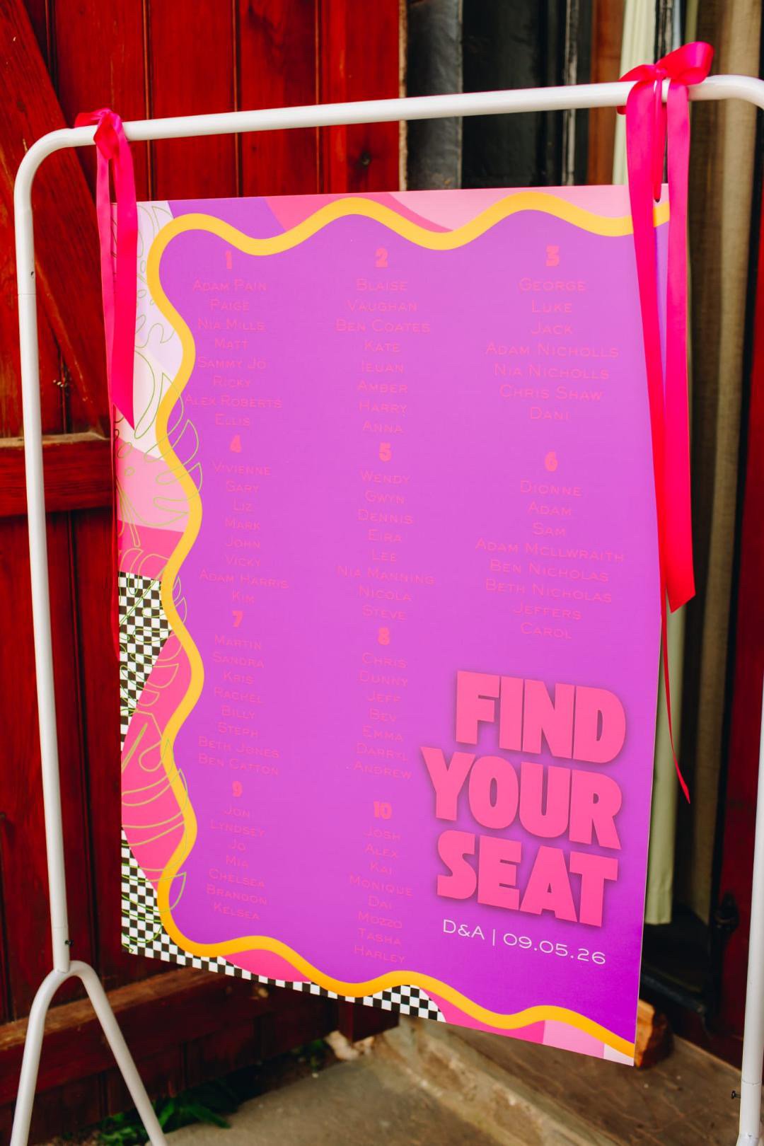 A pink and purple sign with a list of names and numbers, and the phrase 'FIND YOUR SEAT' in large pink letters, with a pink ribbon attached at the top. The sign is on a white stand against a wooden fence, indicating a seating chart for a wedding or e