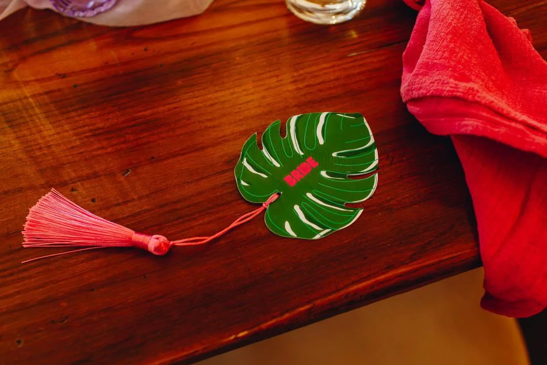 Decorative bookmark shaped like a green monstera leaf with the word "BRAVE" in red, attached to a pink tassel, resting on a wooden table with a red cloth nearby.