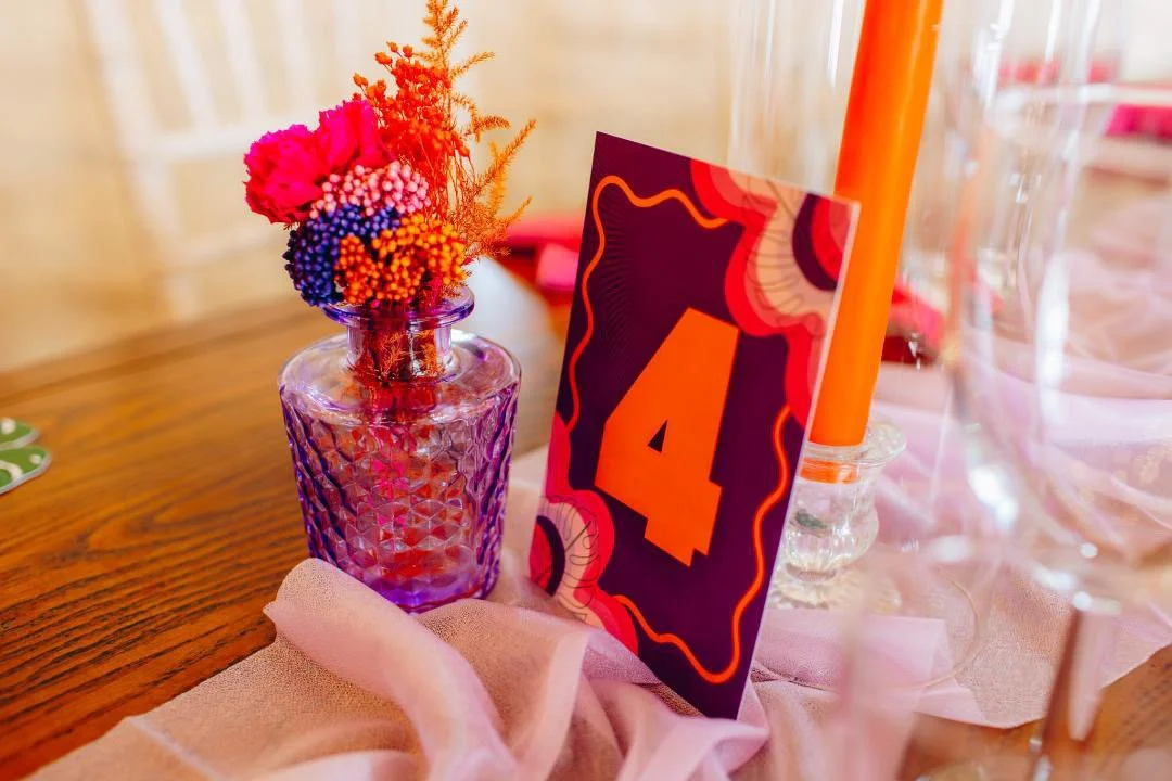 A table arrangement includes a purple glass vase with pink, orange, purple, and red artificial flowers, a table number card with the number 4, a tall orange candle in a clear glass holder, and a pink cloth on a wooden table. With bells on.
