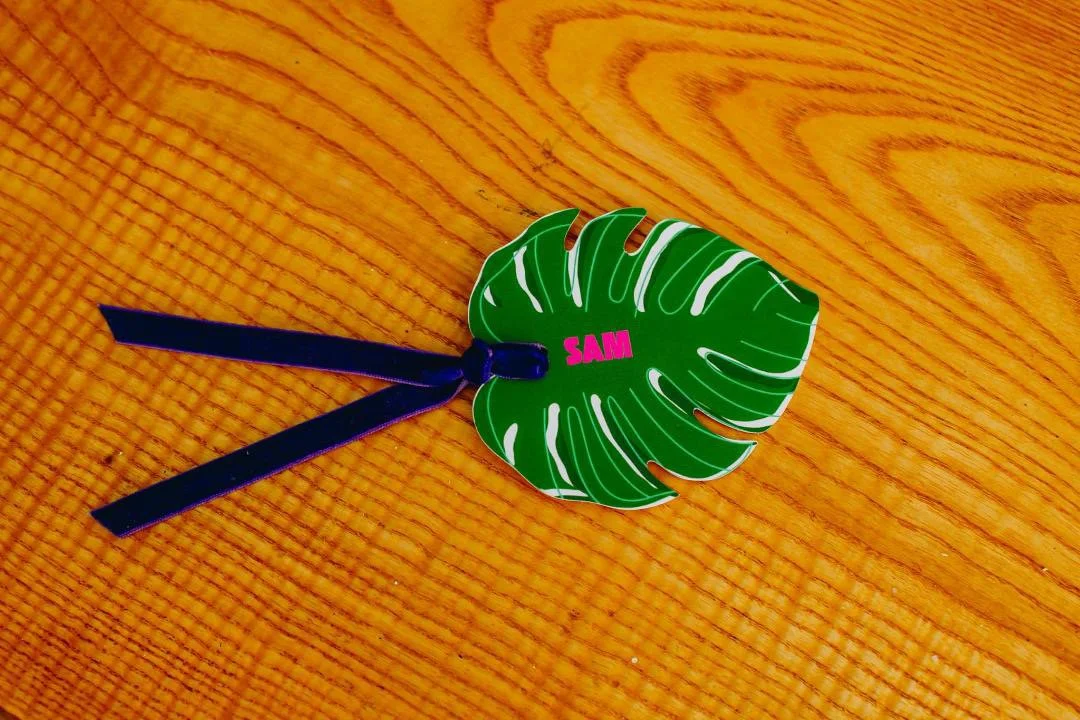 A decorative hair clip resembling a green monstera leaf with white veins and the name 'SAM' written in pink in the center, attached to a black ribbon, placed on a wooden surface.