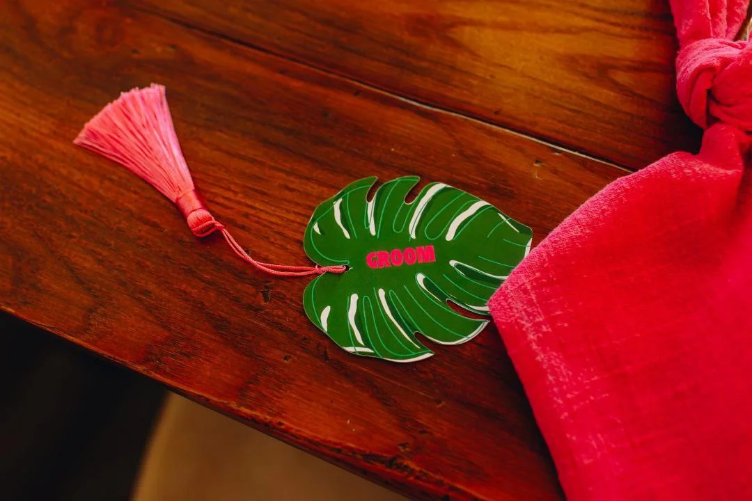 A green decorative item shaped like a monstera leaf with the word 'GROOM' in pink, attached to a pink tassel, resting on a wooden surface next to a pink cloth. Bespoke stationery with tassel embellishments.