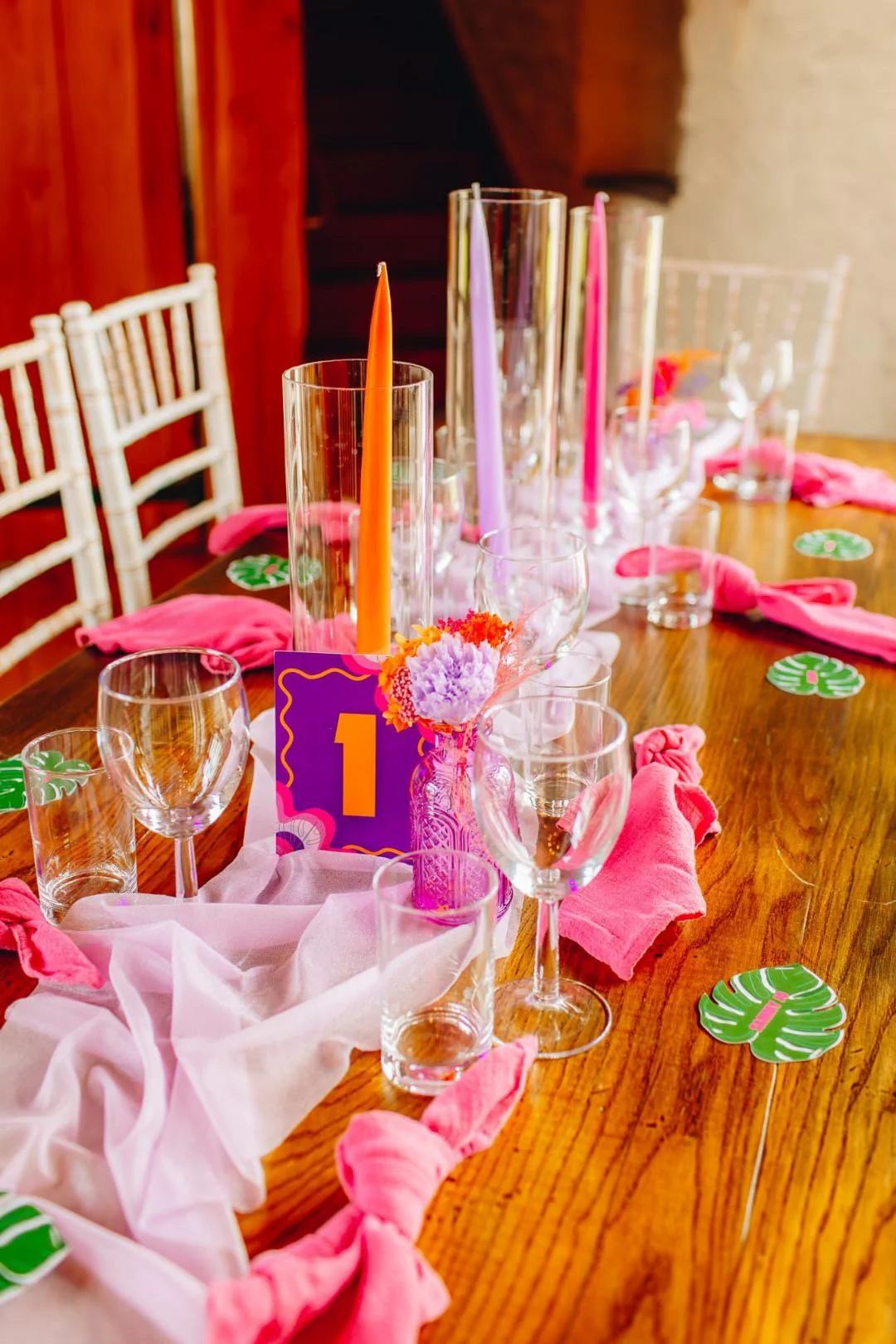 Table decorated for celebration with pink and purple accents, glassware, candles, table number, and pink cloth napkins.