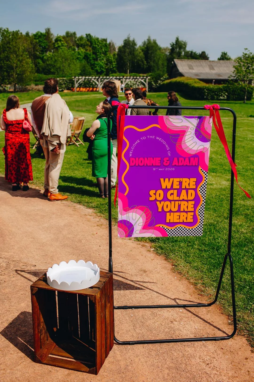 Wedding welcome sign that says 'Welcome to the wedding of Dionne & Adam, 9th May 2026. We're so glad you're here', with a purple background and colorful abstract designs, positioned outdoors on a grassy area. In the background, people are gathered, t