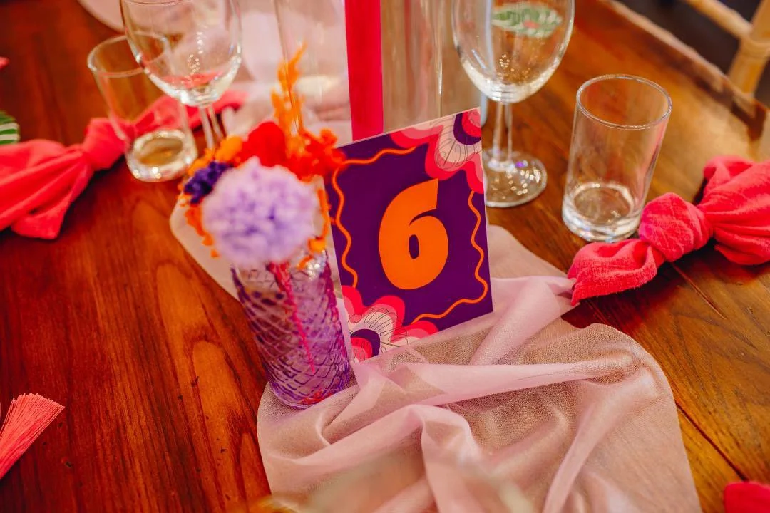 Table setting with clear glasses, a pink table runner, red cloth tied in knots, and a centerpiece with a card displaying the number six.