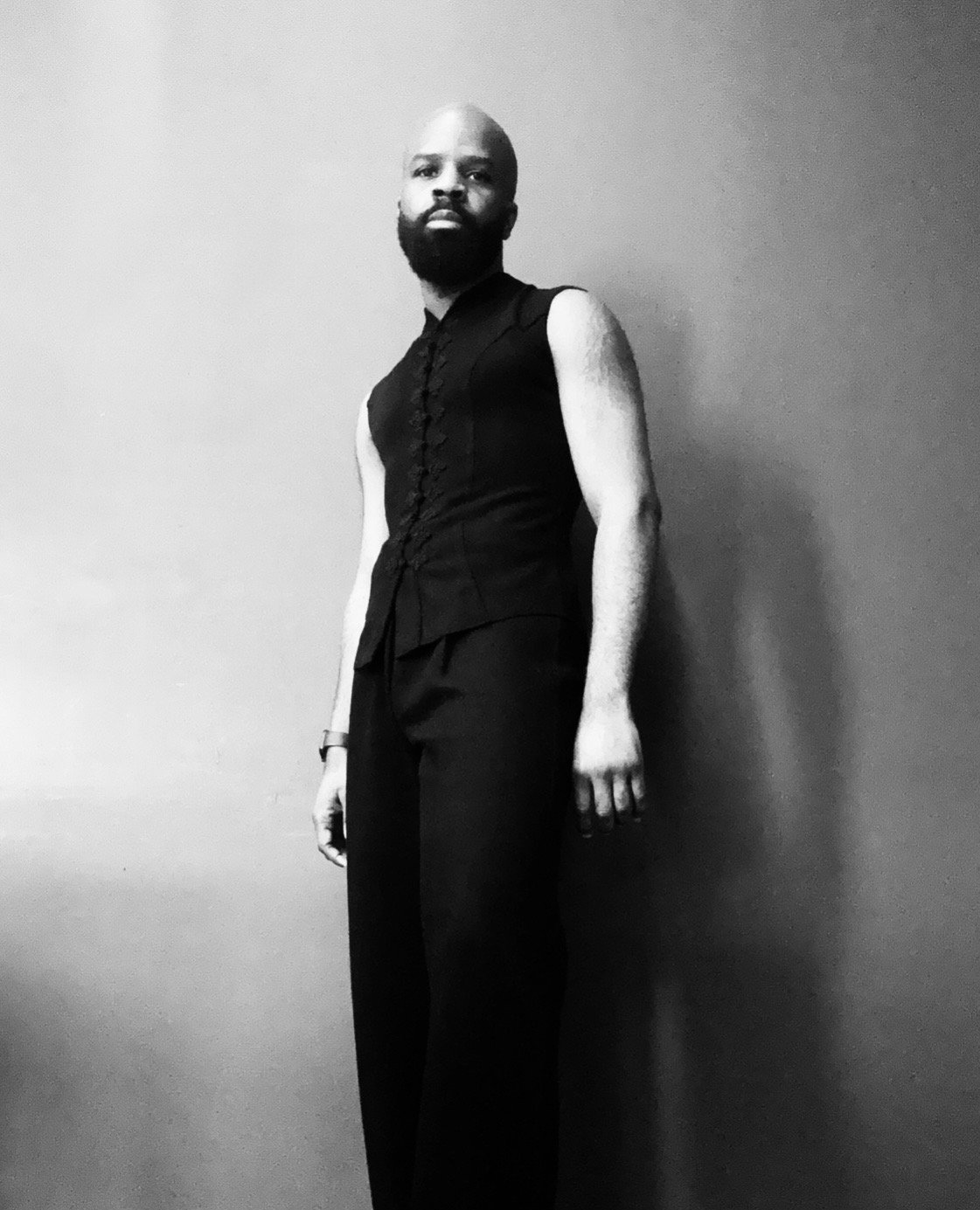 A man with a beard and bald head standing against a plain wall, wearing a sleeveless black top and black pants, captured in black and white.