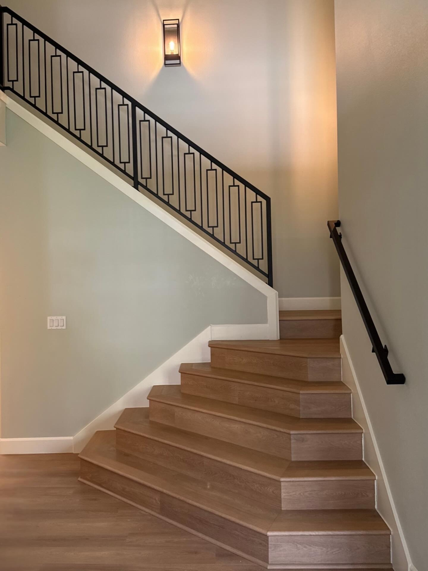 Interior staircase with wooden steps, black metal railing on the upper part, a wall-mounted light fixture, and a wooden handrail on the right side.