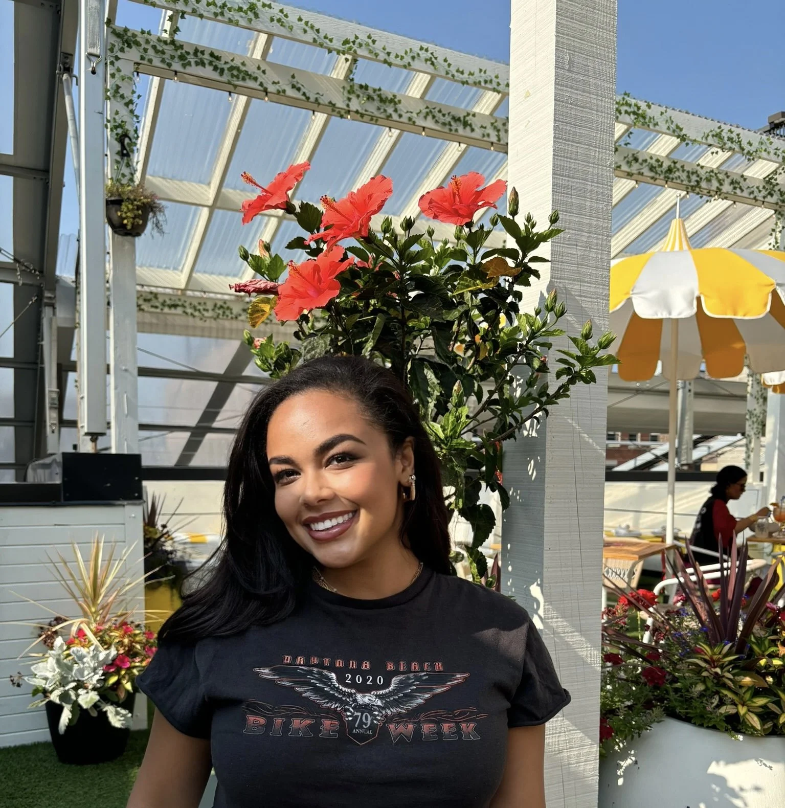 A smiling woman with black hair, wearing a black T-shirt with a graphic that reads 'Daytona Beach 2020 Bike Week,' standing outdoors among colorful flowers and greenery, with a yellow and white striped umbrella and a glass-roofed structure in the background.