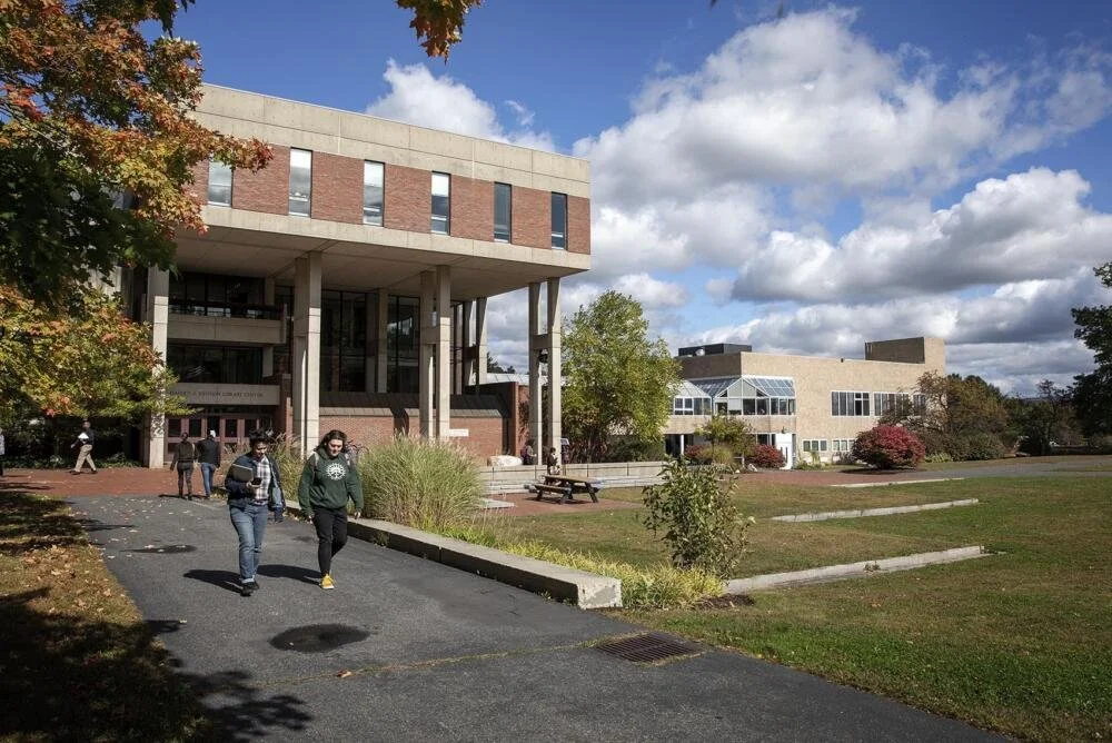 College campus with students walking on a pathway, modern buildings, trees with autumn foliage, and a partly cloudy sky.