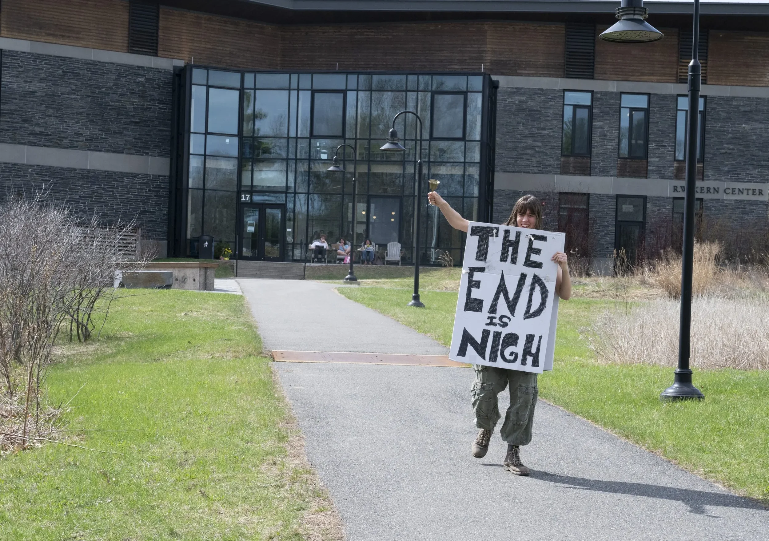 A person holding a sign that reads 'The End is Nigh' walking down a paved pathway outside a modern building with large glass windows, while other people sit on chairs near the entrance.