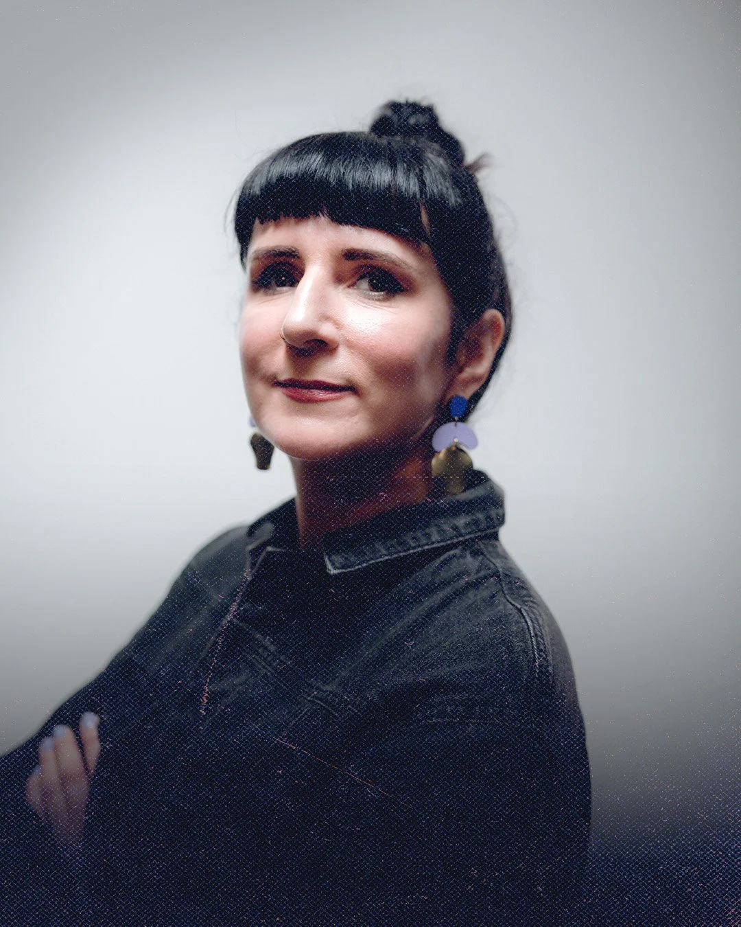 Close-up portrait of a woman with dark hair styled in a bun, wearing colorful earrings, black jacket, and posing with arms crossed against a plain light-colored background.