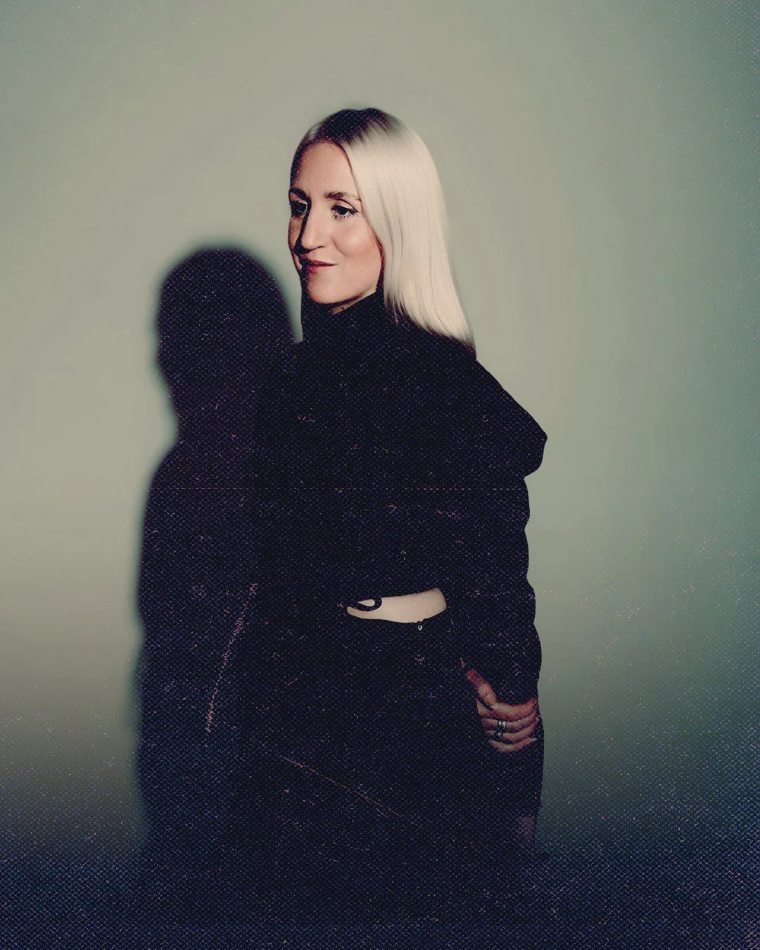 A woman with platinum blonde hair and light skin, wearing a black hoodie, standing against a plain light green background, casting a shadow to her left.