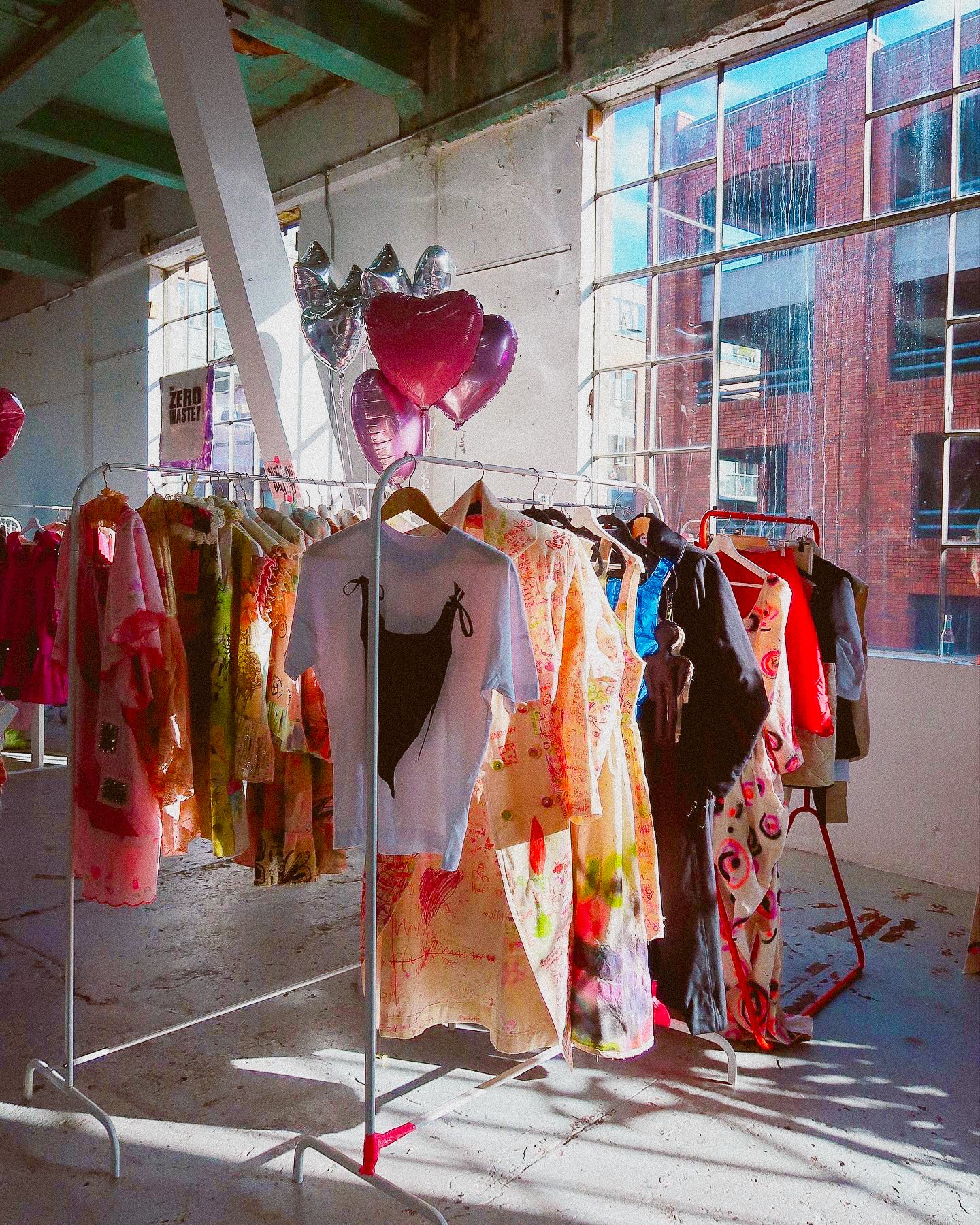 Clothing display with colorful dresses and jackets on racks in an industrial-style room with large windows and balloons.