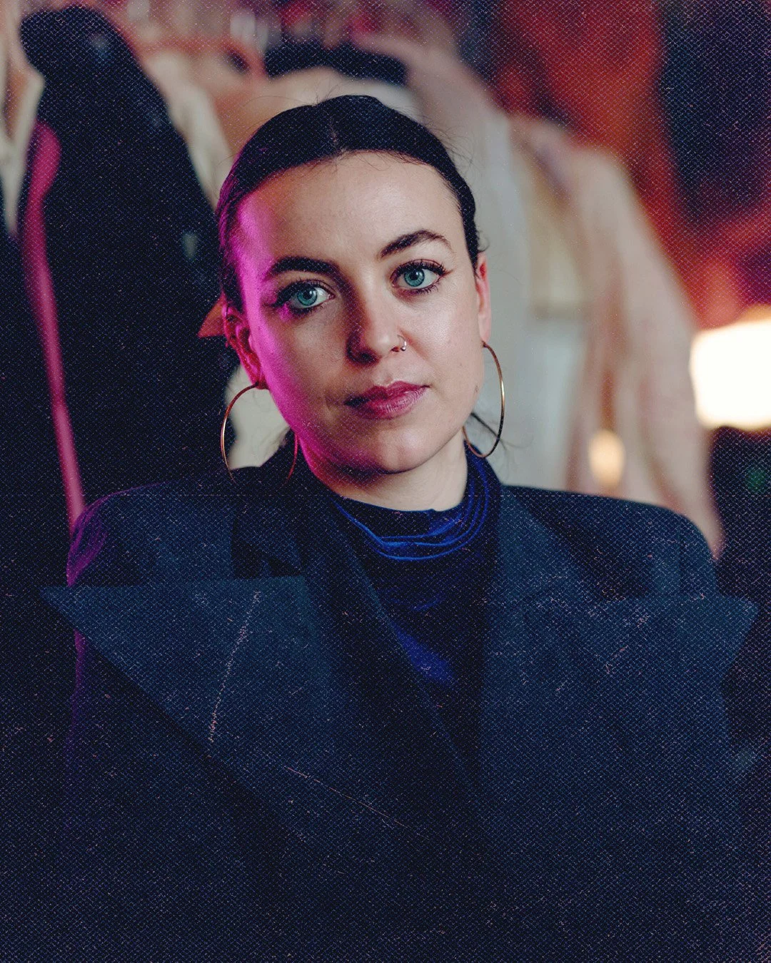 A woman with blue eyes, dark hair, and large hoop earrings gazes into the camera with a confident expression. She wears a black jacket and a dark top with a high collar. The background is blurred, showing other people at a social event.