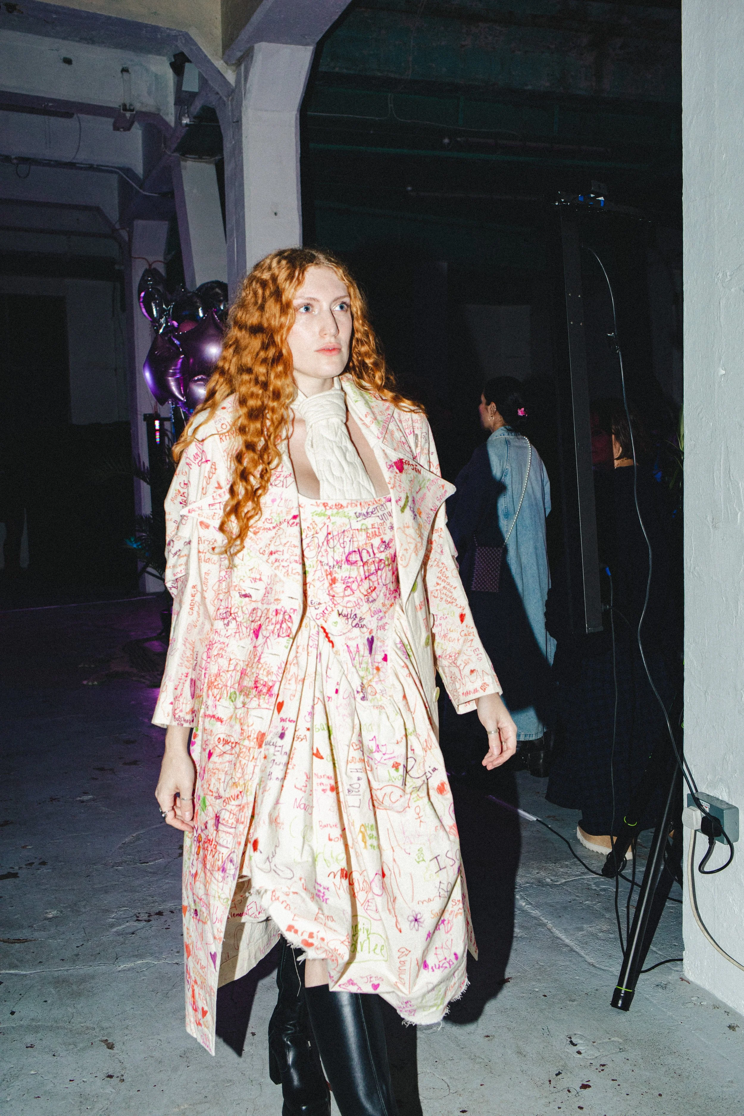 A woman with long red curly hair wearing a cream-colored dress with pink and red signatures written all over it. She is also wearing black boots. In the background, there are purple balloons and other people standing in a room with a concrete floor and white wall.