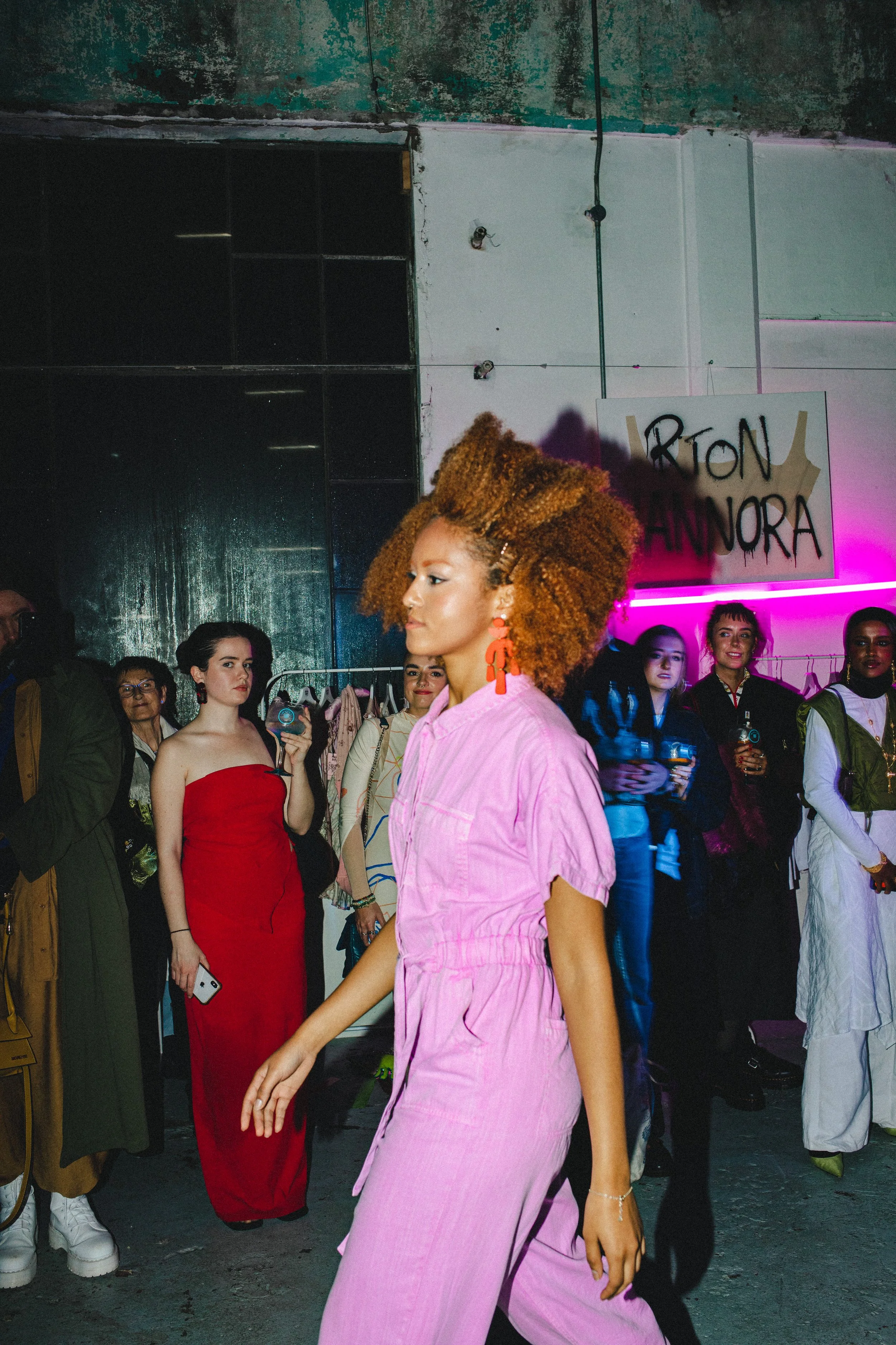 A woman with curly, orange hair wearing a pink jumpsuit walks past a group of people at what appears to be an indoor event or fashion show. The background features an industrial space with exposed walls, some neon lighting, and a sign that partially reads 'RTON HANORA.' The audience includes women dressed in stylish outfits, some holding phones or drinks.