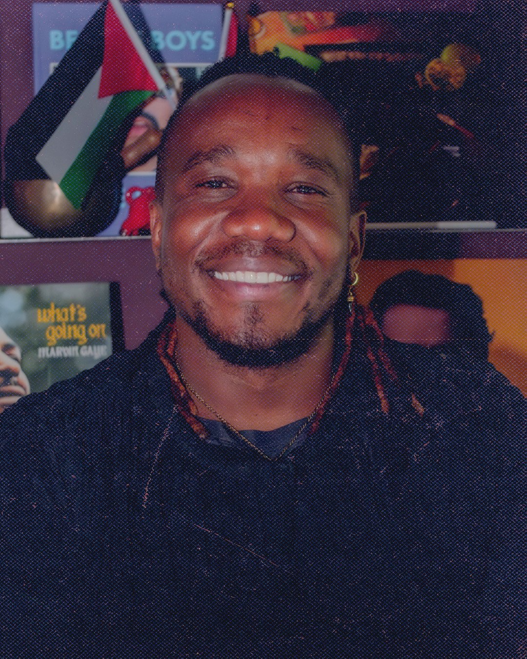 A smiling man with a beard and braids, wearing a black shirt, in front of a background with various posters and flags.