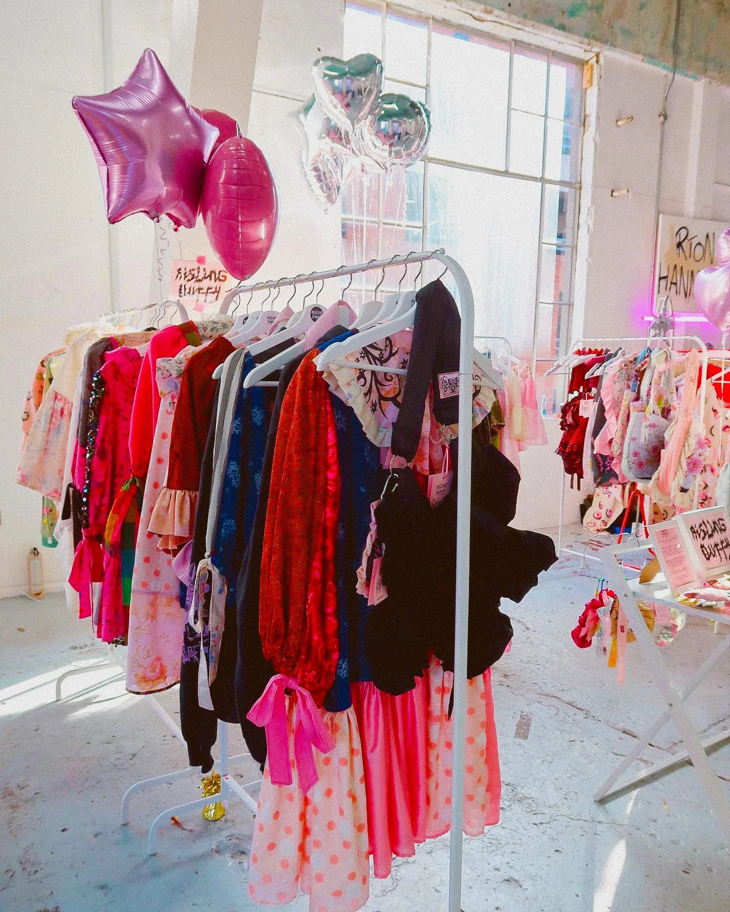 A clothing display with pink, red, blue, black, and white dresses and tops, topped with pink and silver balloons in a bright, industrial-style space with large windows.
