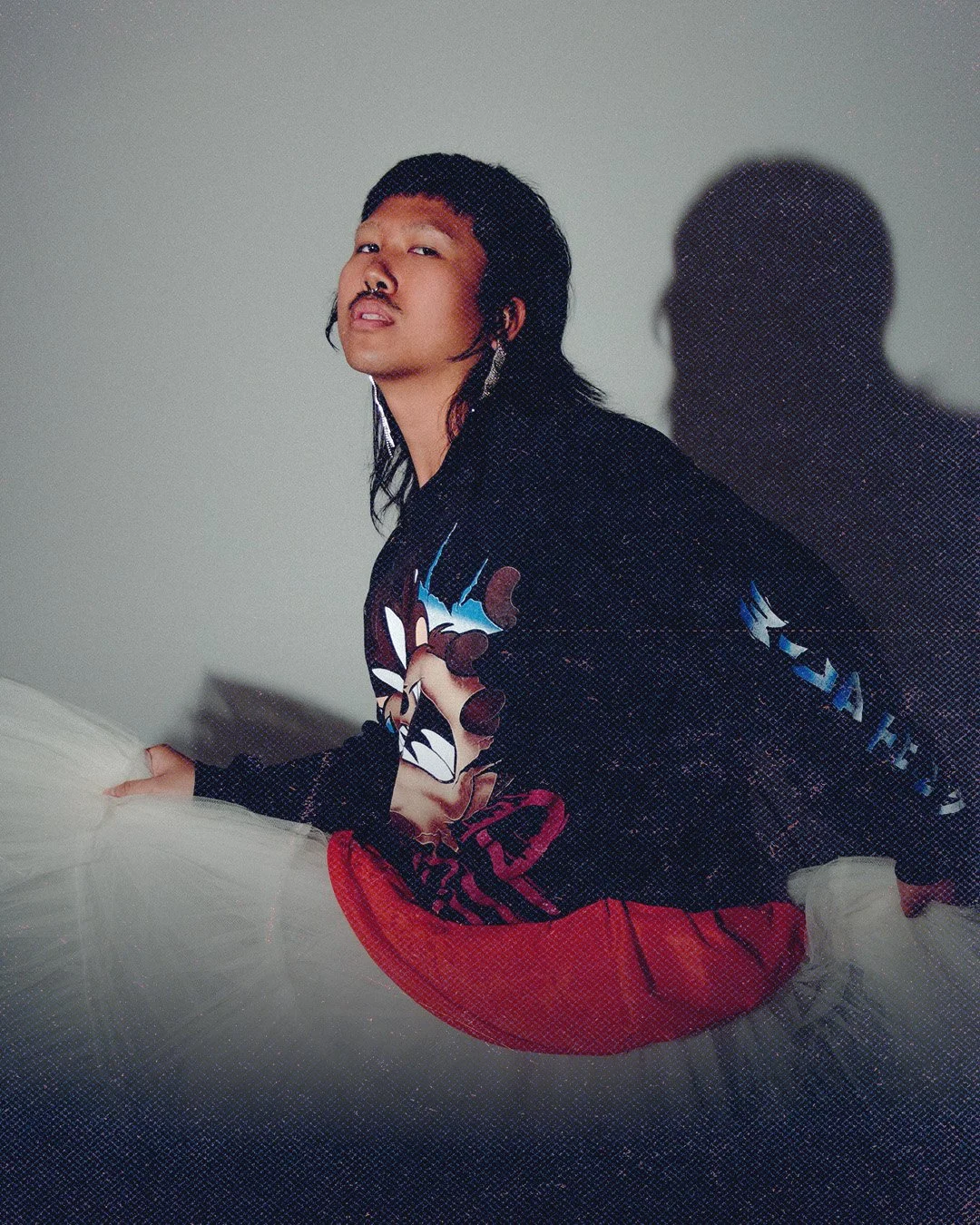 A young man with long black hair and a mustache, wearing a black graphic sweatshirt and red pants, is sitting on a bed with a light grey headboard. His shadow is cast on the plain light grey wall behind him. The photo has a slightly textured overlay.
