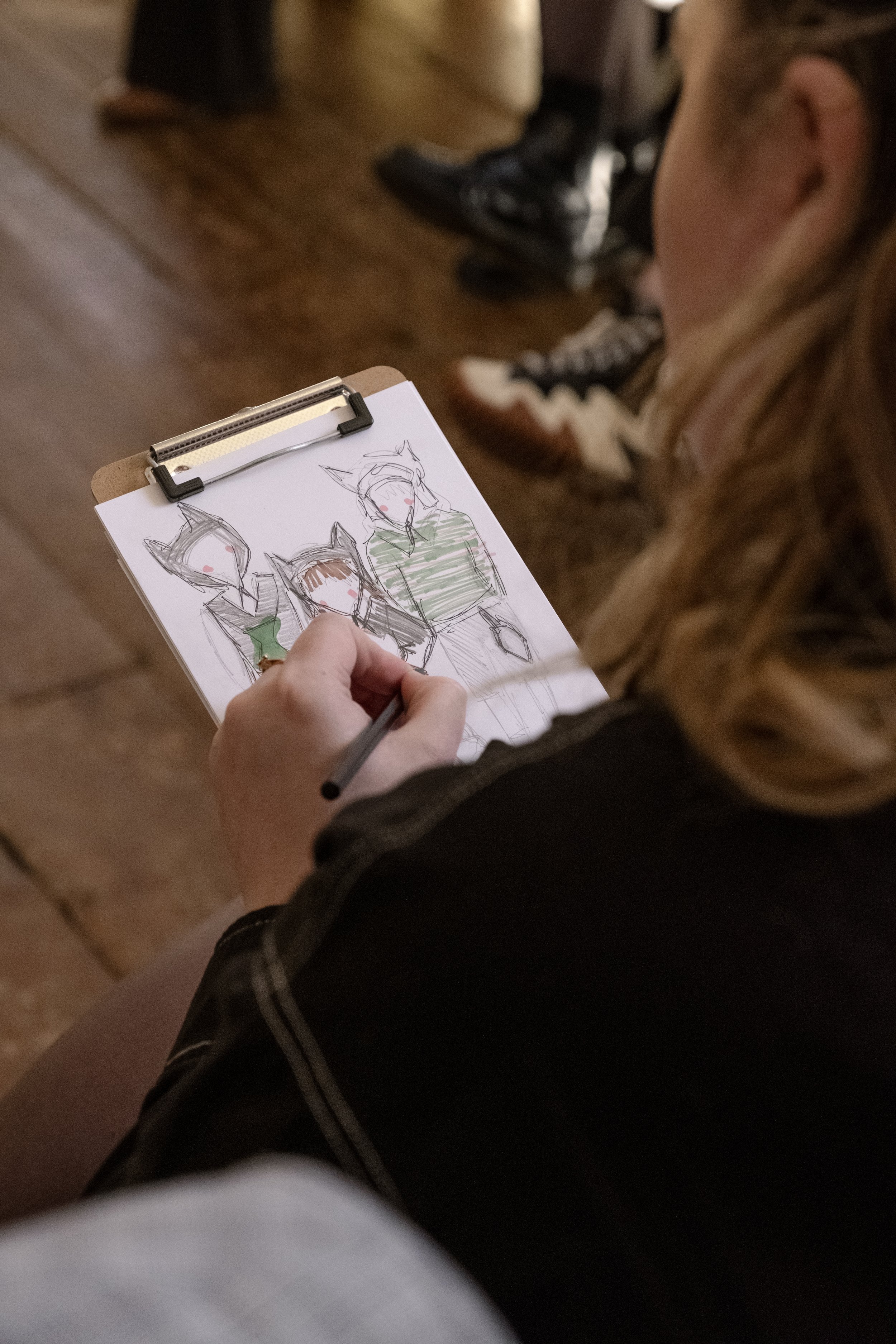 A person is drawing a sketch of three characters with animal-like features on a clipboard in a room with a wooden floor. The drawing includes two characters with fox-like ears and one with dog-like ears, and they have blush marks on their cheeks.