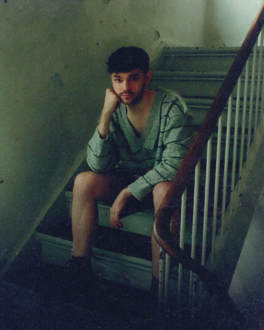 Young man sitting on a staircase, resting his head on his hand, wearing a striped sweater and shorts, in a dimly lit indoor setting.