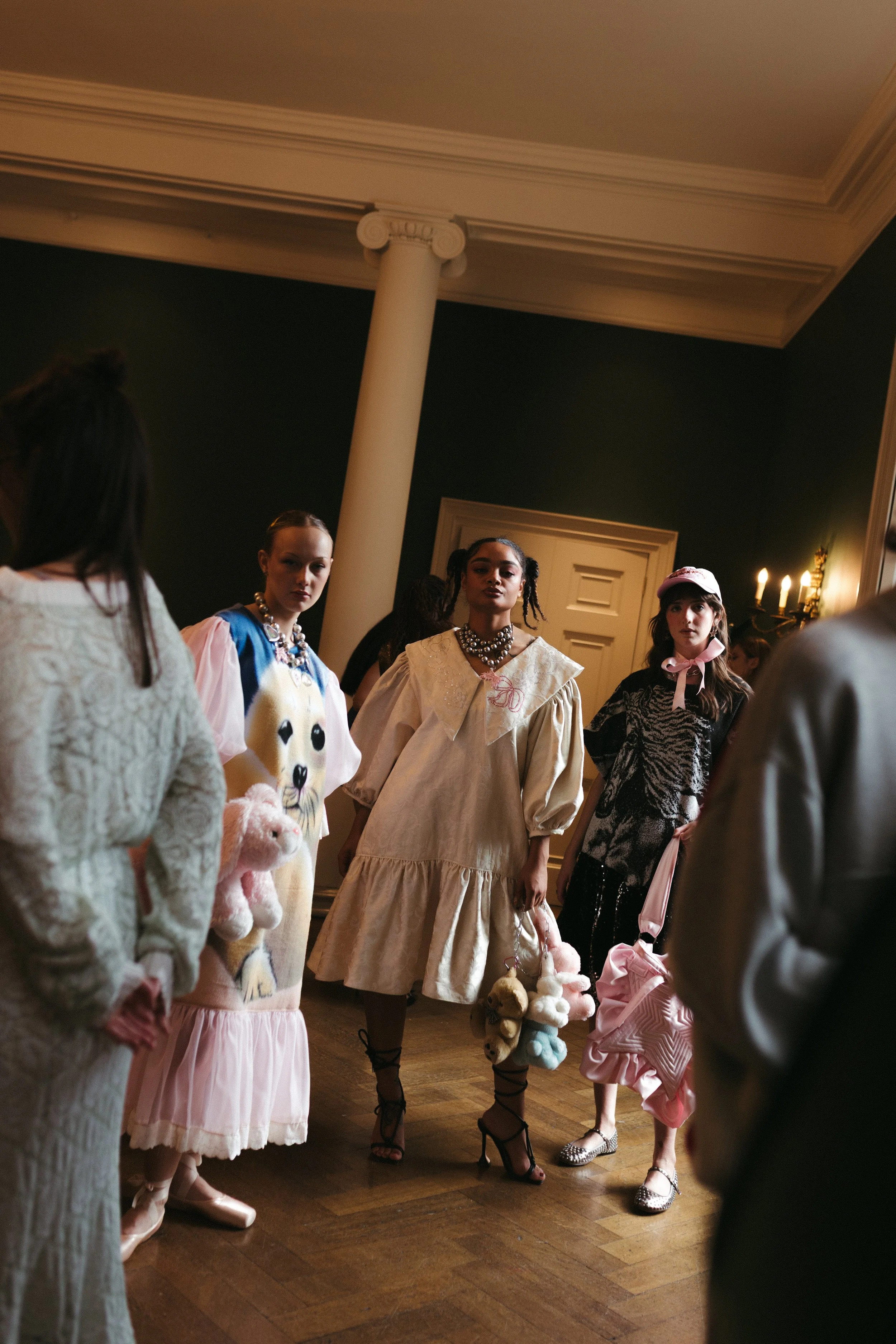 A group of young women dressed in fashionable, eclectic clothing, standing in a room with wooden floors and dark walls, illuminated by warm candlelight, engaged in conversation or awaiting an event.
