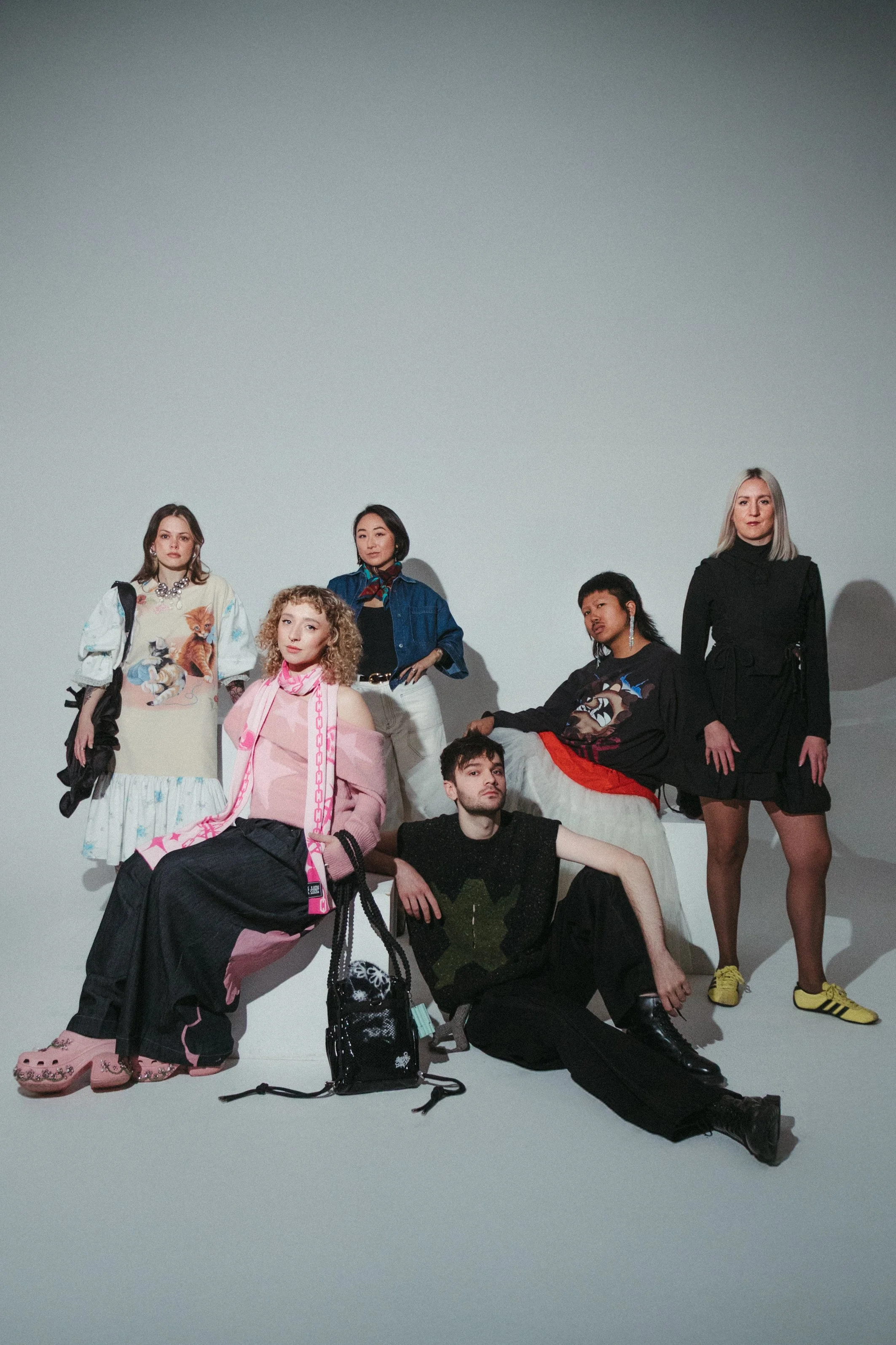 A diverse group of seven young people posing in fashion outfits against a plain white background. Some are standing, others sitting or kneeling. They are accessorized with jewelry, bags, and colorful sneakers, giving a modern, edgy vibe.