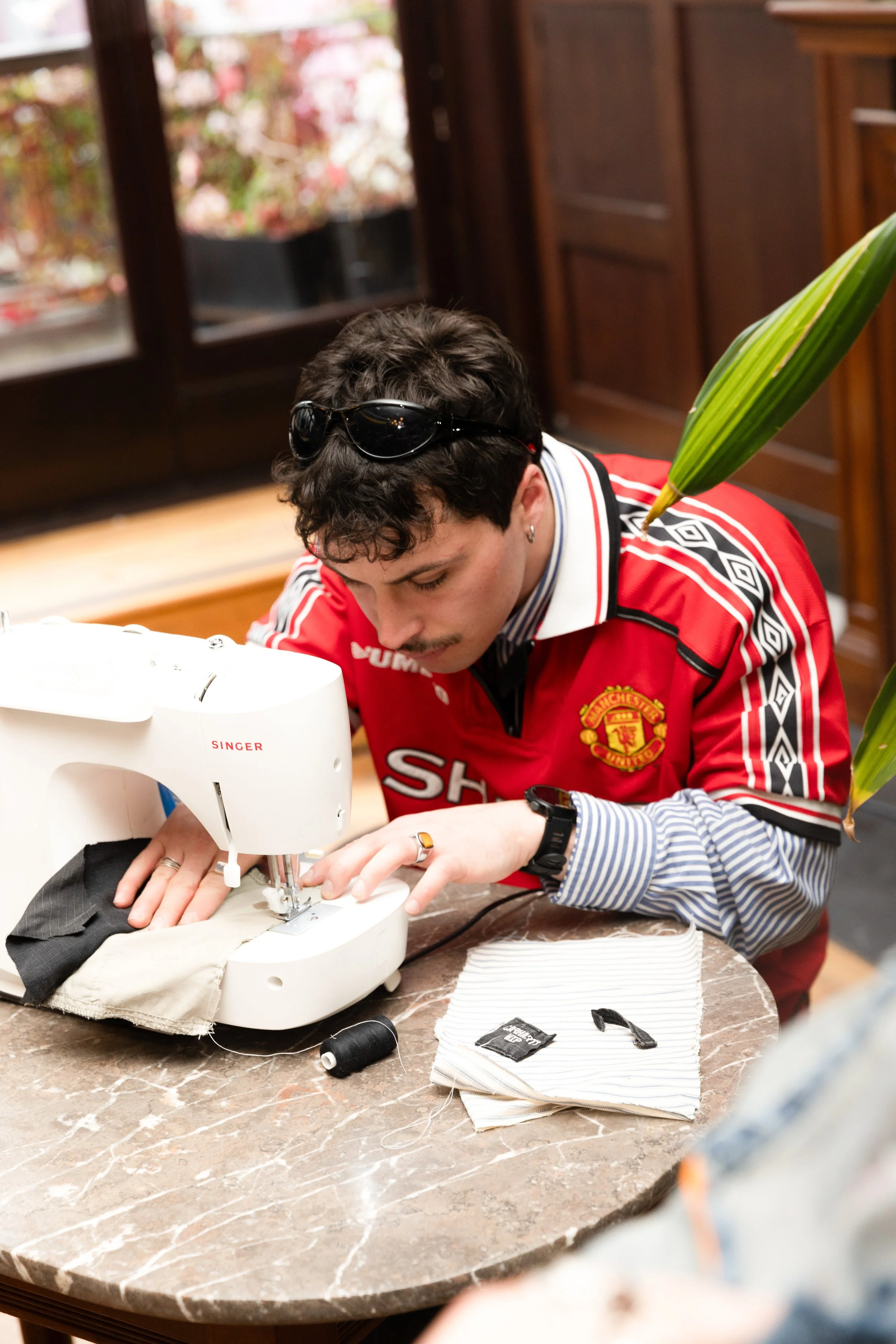 Person sewing fabric on a white sewing machine at a marble table. The person has dark hair, sunglasses on head, and a Manchester United jersey. Sewing supplies and fabric are on the table, with a window and potted plant in the background.