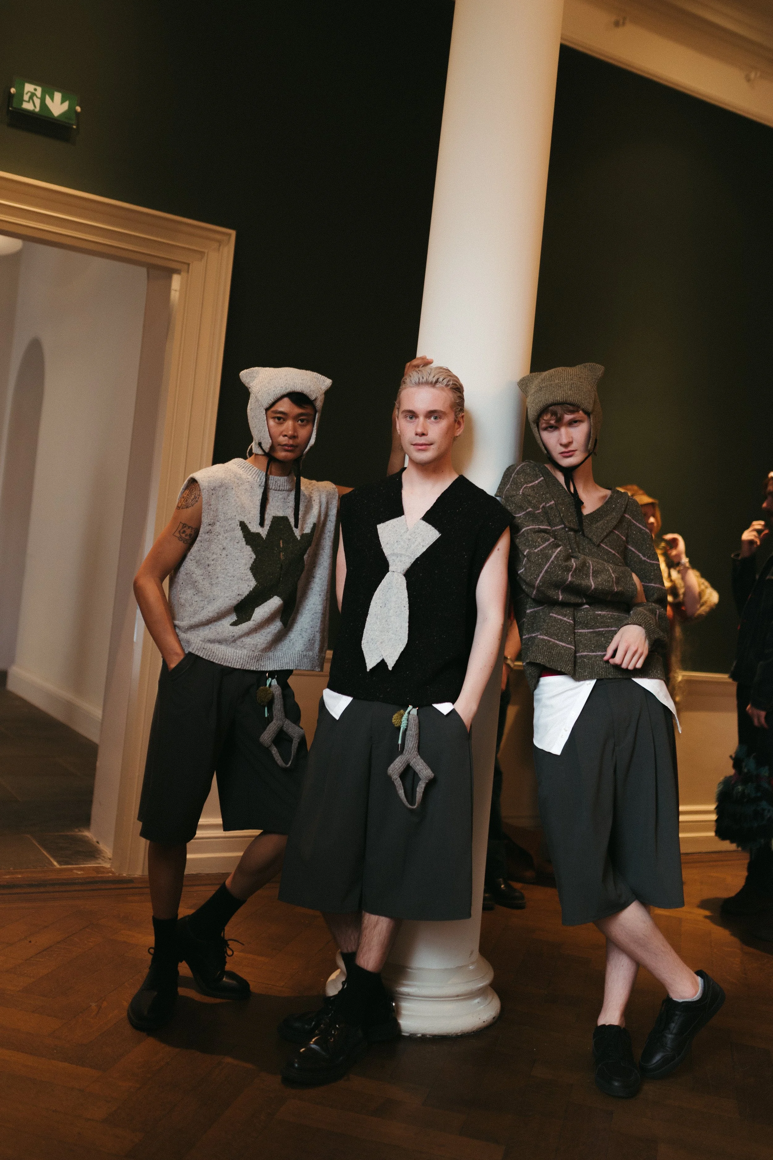 Three young women posing indoors near a white pillar, wearing casual and edgy clothing, with two wearing knit hats with ear flaps. One woman is wearing a sleeveless sweater with a graphic design, another a sleeveless black top with a large tie graphic, and the third a loose brown checked jacket. They are standing on a wooden floor in front of a dark green wall and a doorway.