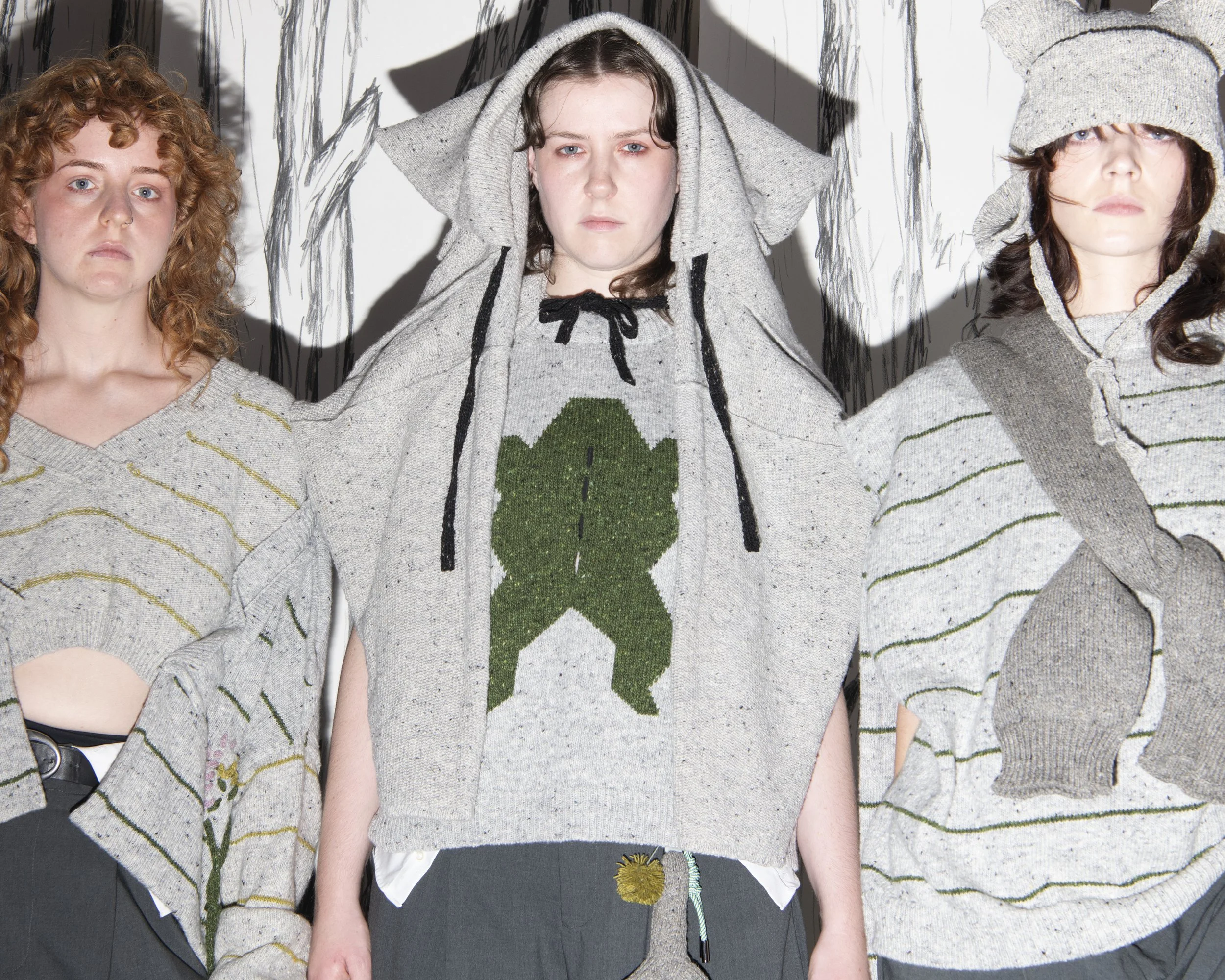 Three young women with different hair colors and styles, wearing cozy sweaters. The woman in the middle is pulling a hood over her head, while the other two have their eyes visible, with one wearing a knit hat. The background features a white wall with black line illustrations of trees.