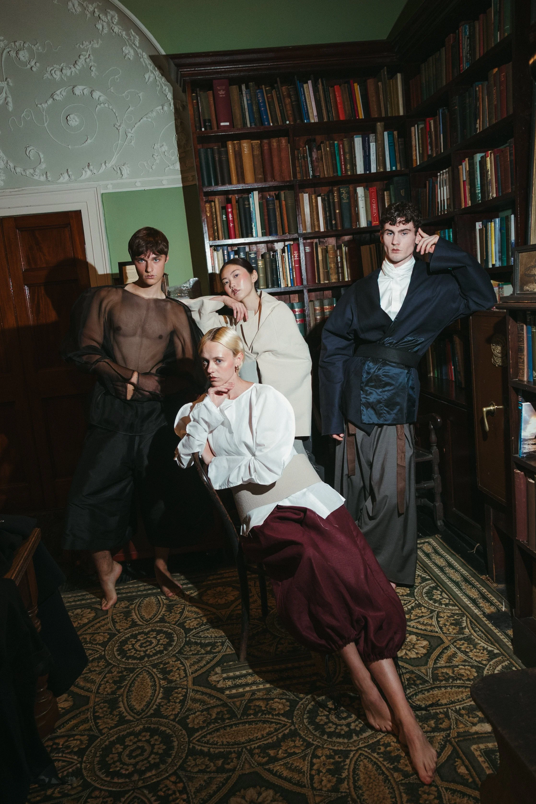 Four models in vintage-inspired clothing pose in a library with bookshelves and ornate ceiling detailing.